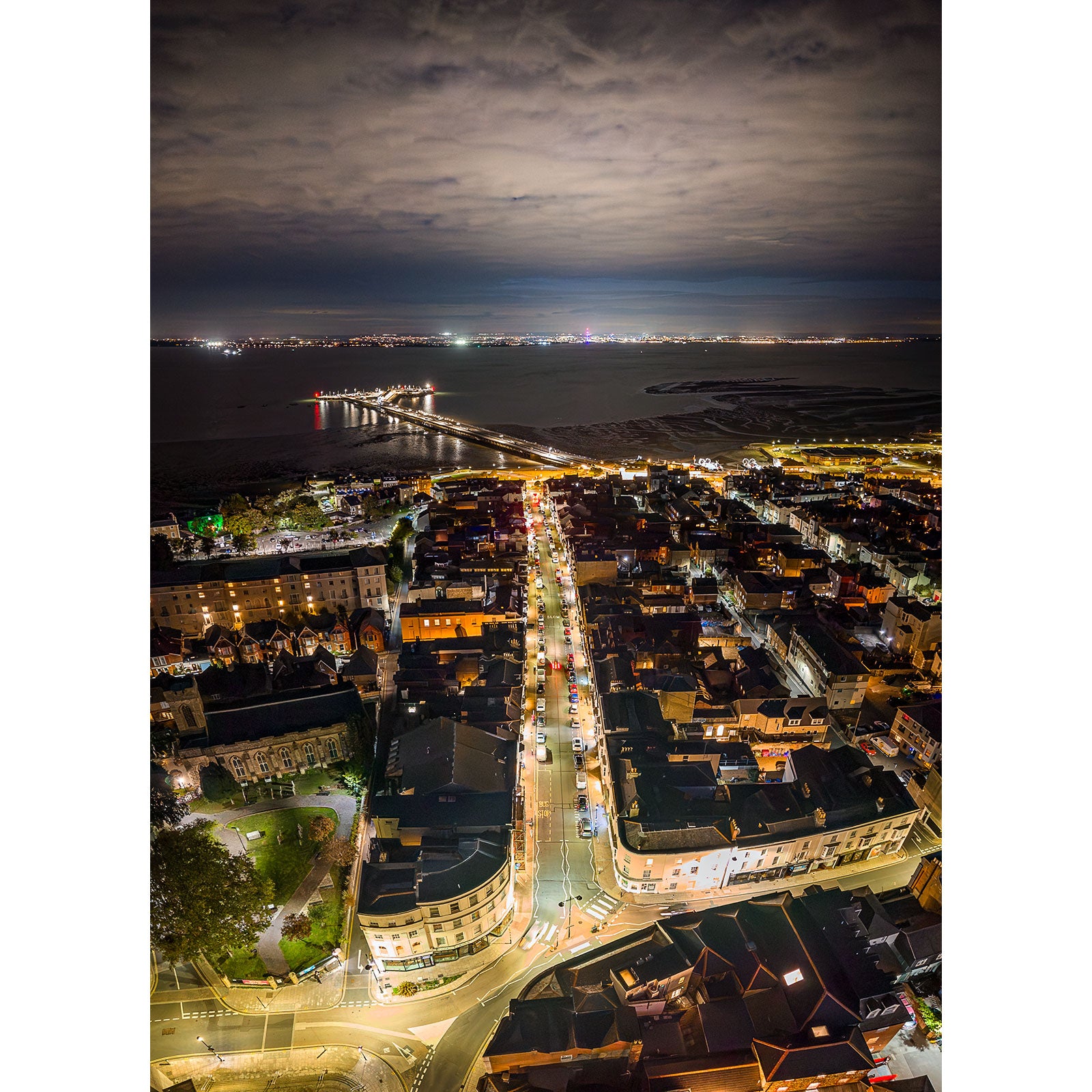 Ryde at Night" by Available Light Photography captures an aerial night view of a coastal city, featuring illuminated streets, a long pier reaching into the sea, and shimmering lights reflecting over the water beneath a cloudy sky.