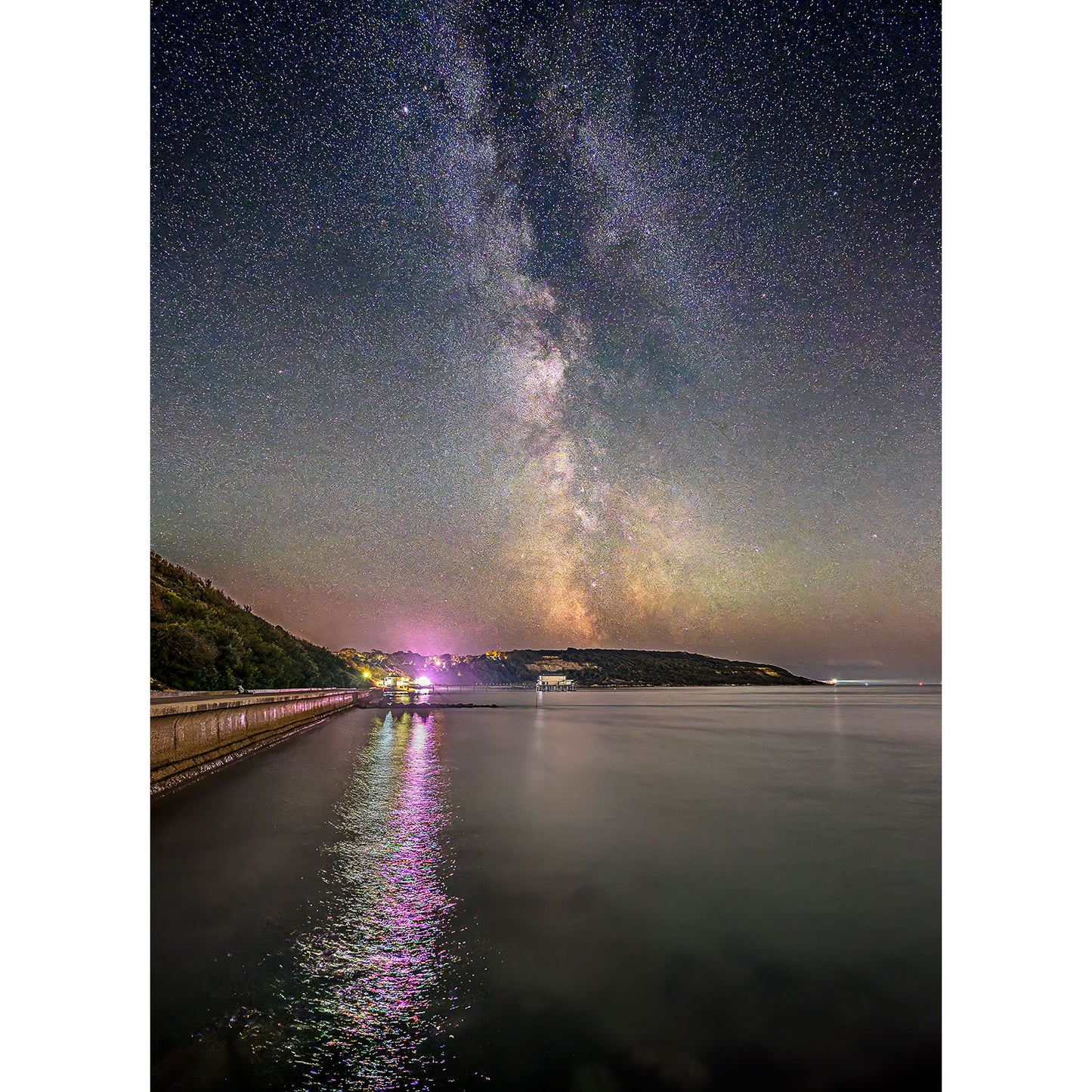 The Milky Way, Totland by Available Light Photography captures the Milky Way spanning the night sky above a tranquil coastline, with colorful lights reflecting and shimmering on the water.