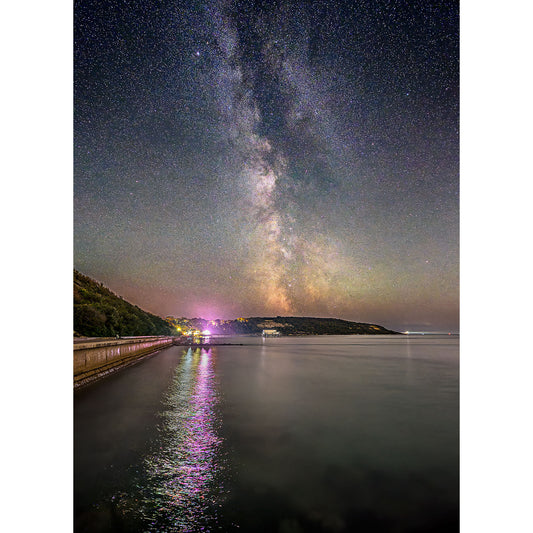 The Milky Way, Totland by Available Light Photography captures the Milky Way spanning the night sky above a tranquil coastline, with colorful lights reflecting and shimmering on the water.