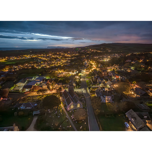 Brighstone at Night" by Available Light Photography captures an aerial view of a small town at dusk, with glowing streetlights illuminating homes, winding roads, and a church among rolling hills under a cloudy sky.