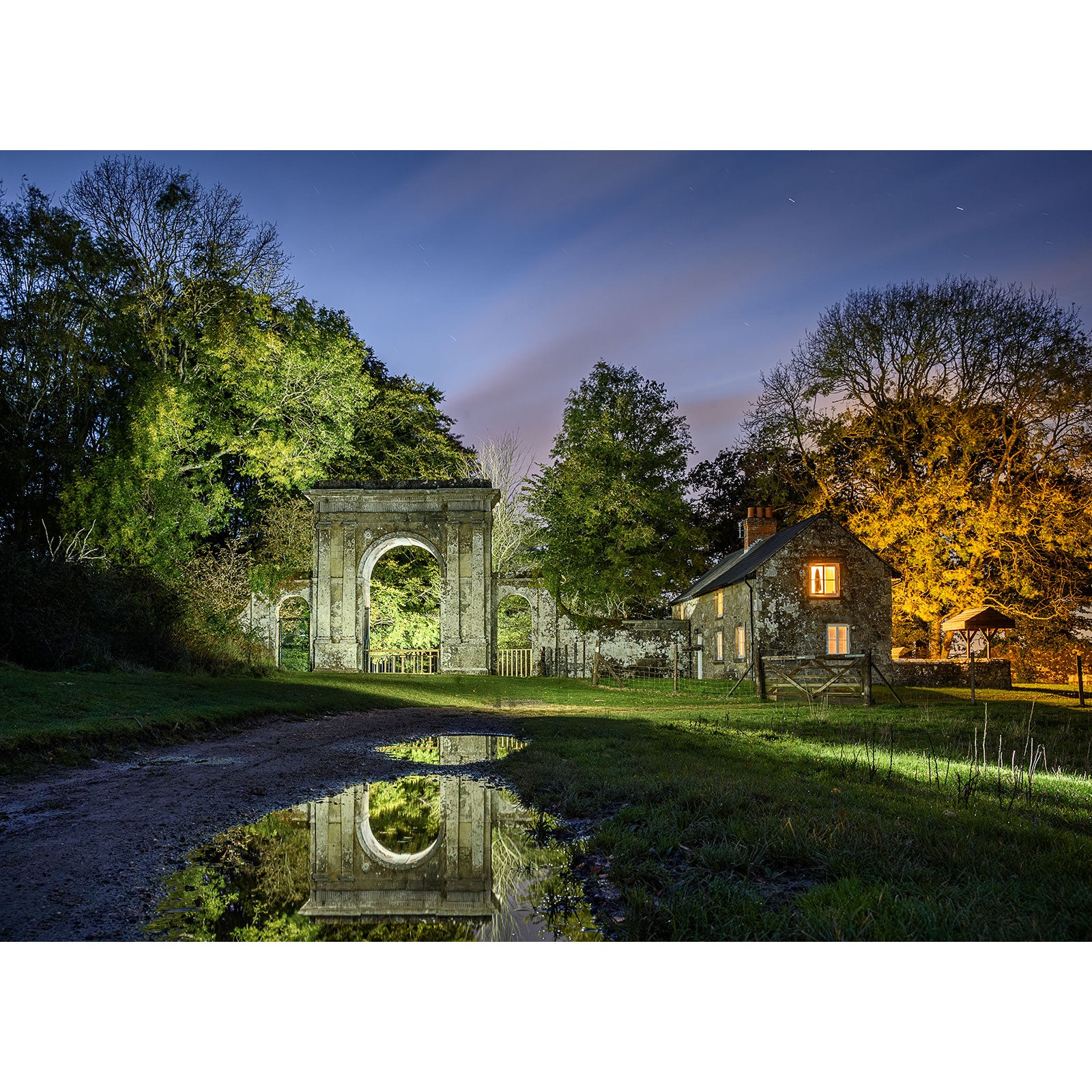 Freemantle Gate, Godshill by Available Light Photography features a stone archway and a small house with glowing windows beside a dirt path, reflected in a puddle and surrounded by trees at dusk.