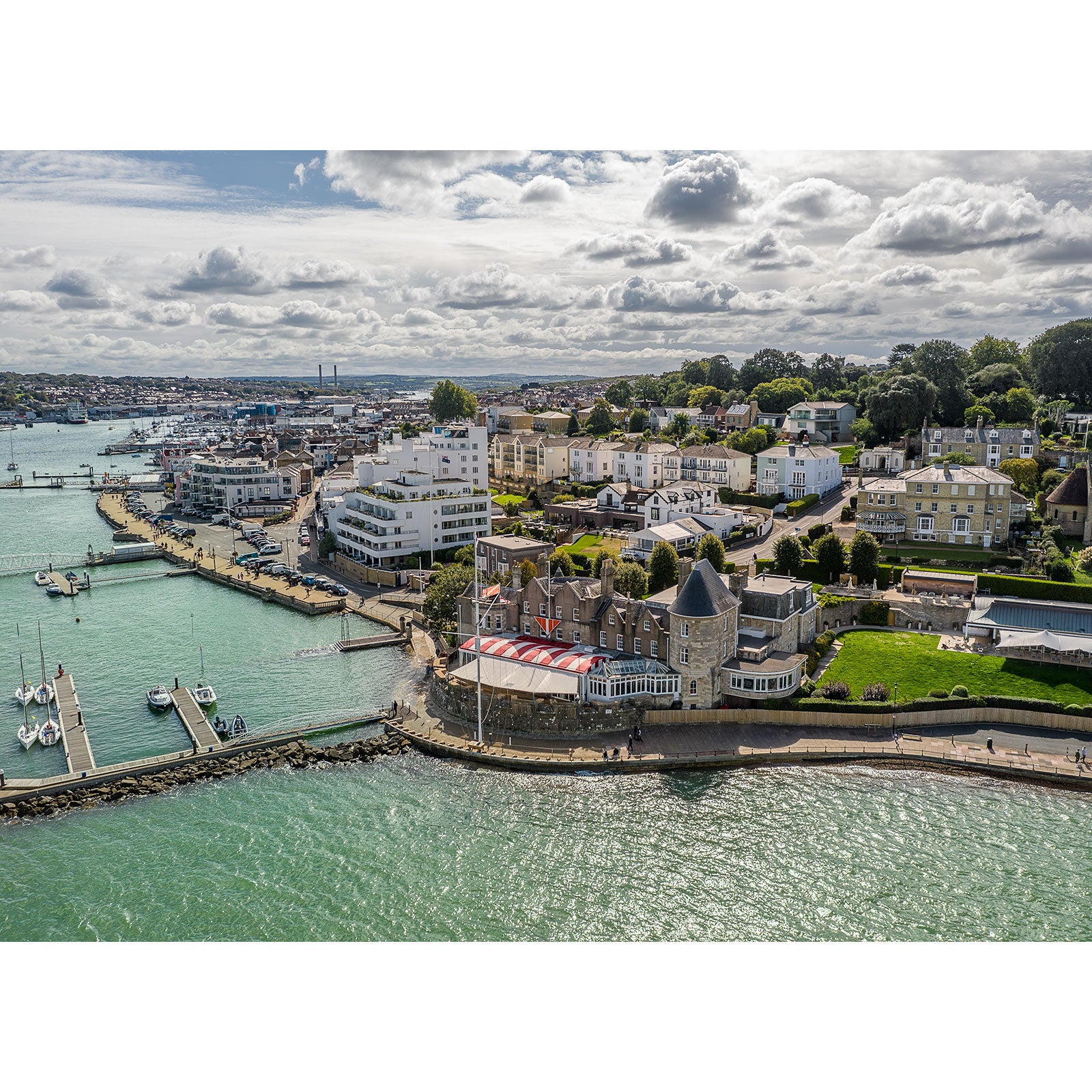 Aerial view of The Royal Yacht Squadron, Cowes by Available Light Photography shows vibrant marinas, waterfront buildings, and sleek boats docked along the shoreline beneath a partly cloudy sky.