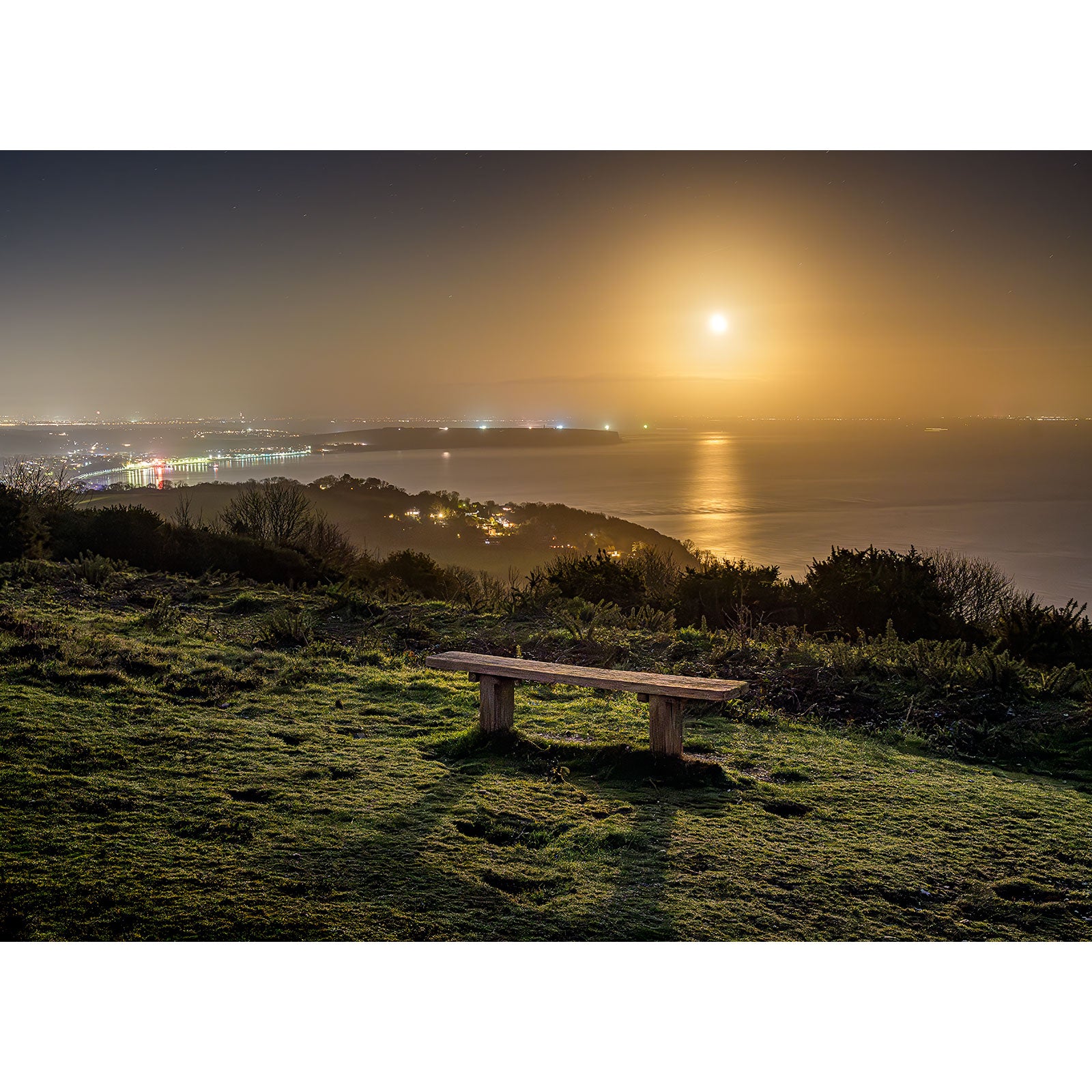 Sandown Bay by Moonlight" by Available Light Photography captures a wooden bench overlooking the tranquil coastal town at night, as moonlight reflects off the sea and gently illuminates the grassy hillside.