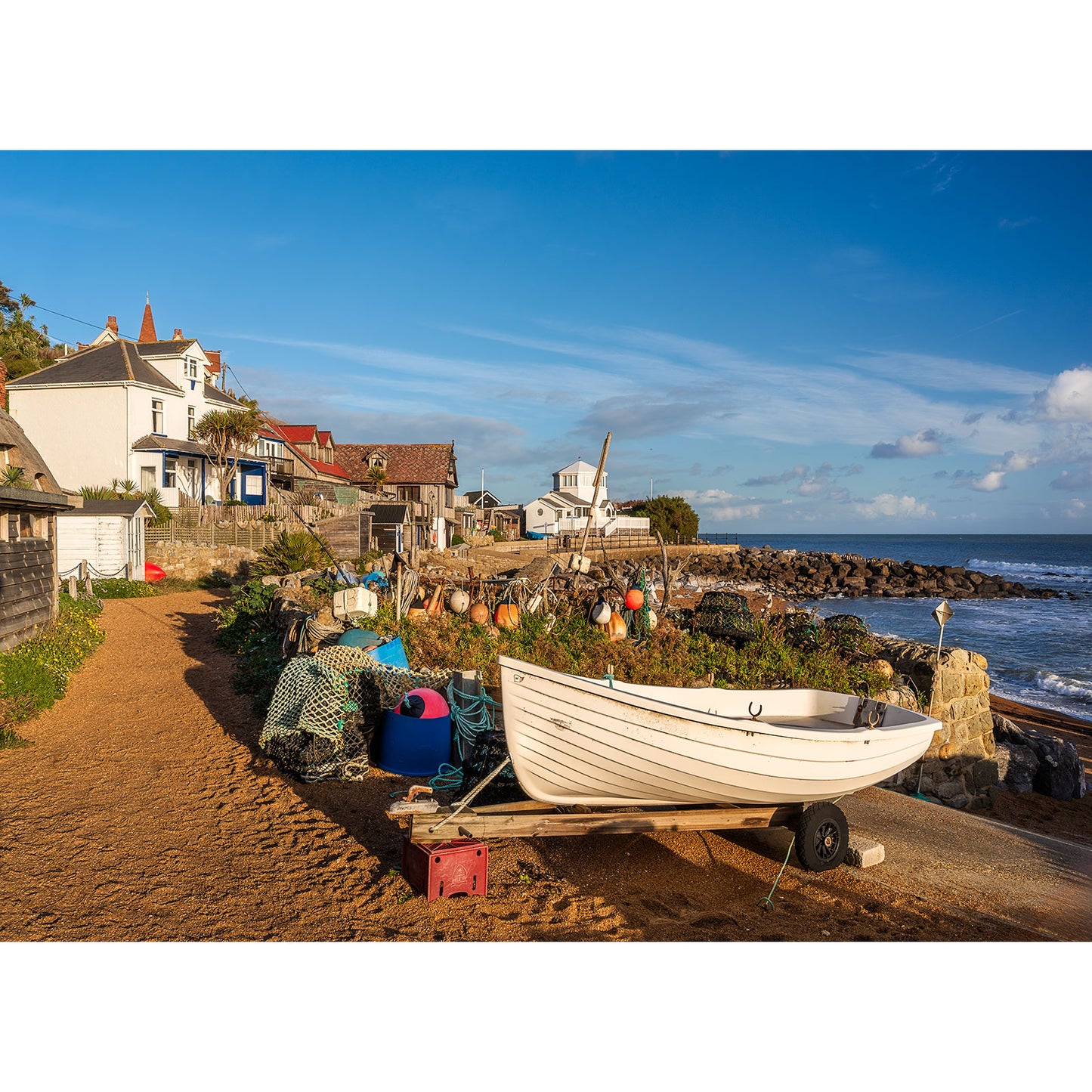 Steephill Cove by Available Light Photography features a small white boat on a sandy path near fishing nets and gear, with coastal houses and the sea in the background under a clear blue sky.