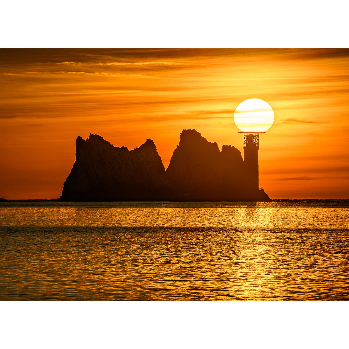 The Needles by Available Light Photography features a lighthouse silhouetted against an orange sunset, the sun appearing to rest atop the beacon near rugged sea stacks.
