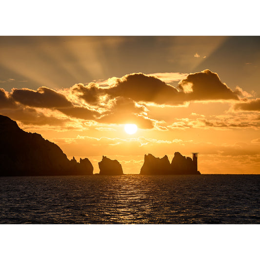 As the sun sets behind The Needles’ jagged sea stacks and cliffs, golden light bathes the tranquil ocean, with rays streaming through scattered evening clouds. Photograph by Available Light Photography.