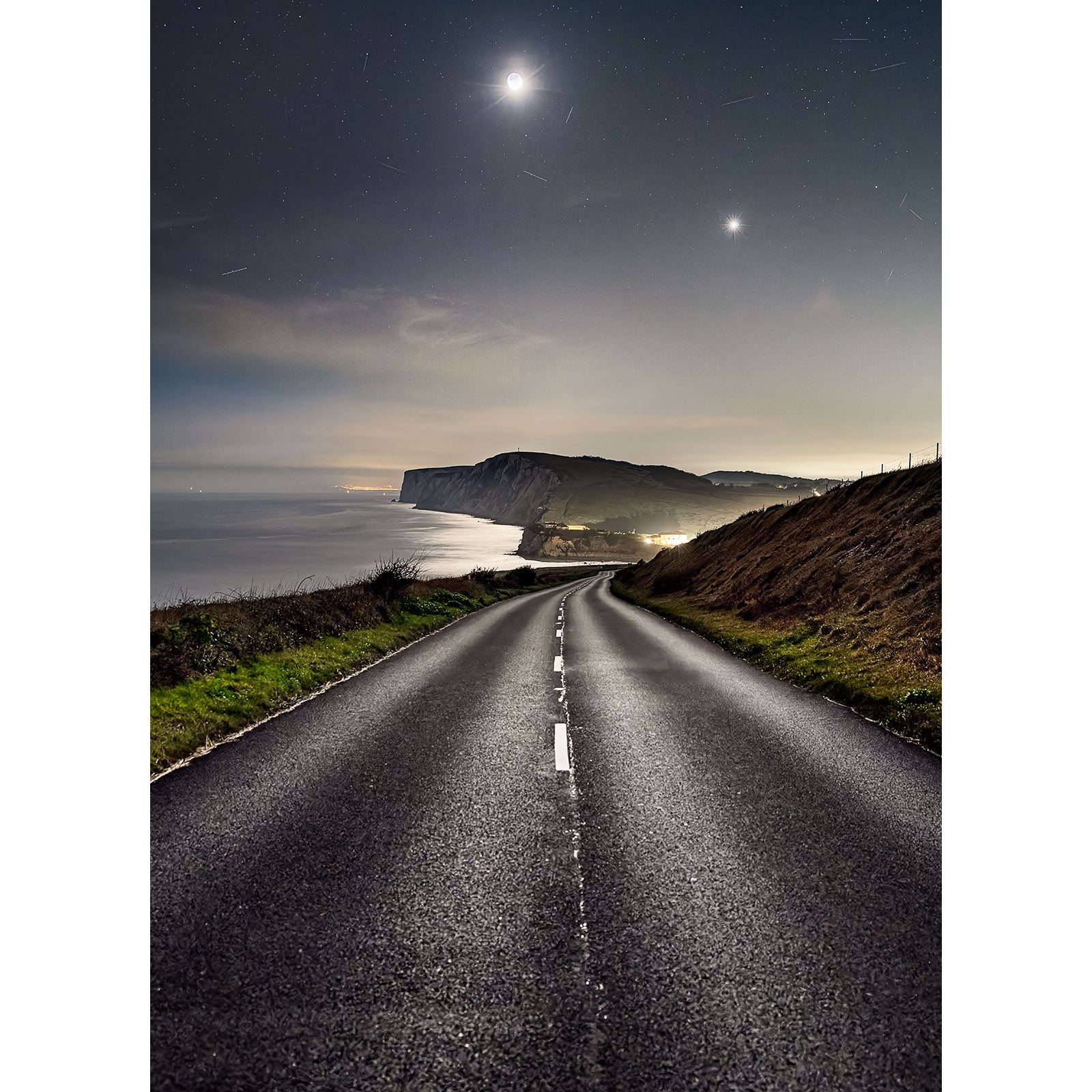 A paved road descends toward the coast at night under cliffs and distant lights, with the crescent moon and Venus shining above. Crescent Moon & Venus, Tennyson Down by Available Light Photography captures this stunning scene.