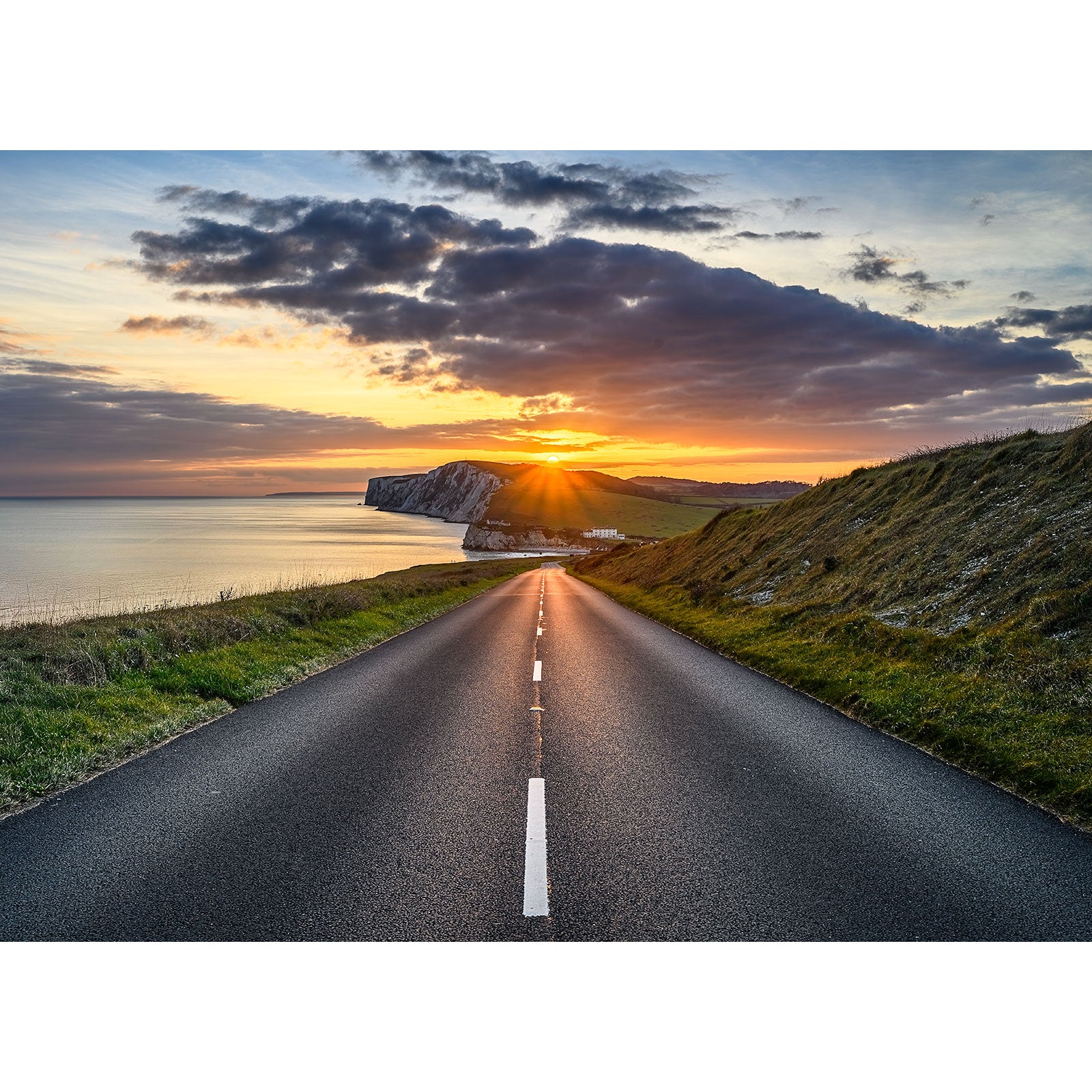 A paved road leads to Tennyson Down’s cliffs and ocean, illuminated by a dramatic sunset sky with clouds, captured by Available Light Photography.