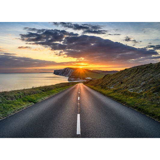 A paved road leads to Tennyson Down’s cliffs and ocean, illuminated by a dramatic sunset sky with clouds, captured by Available Light Photography.