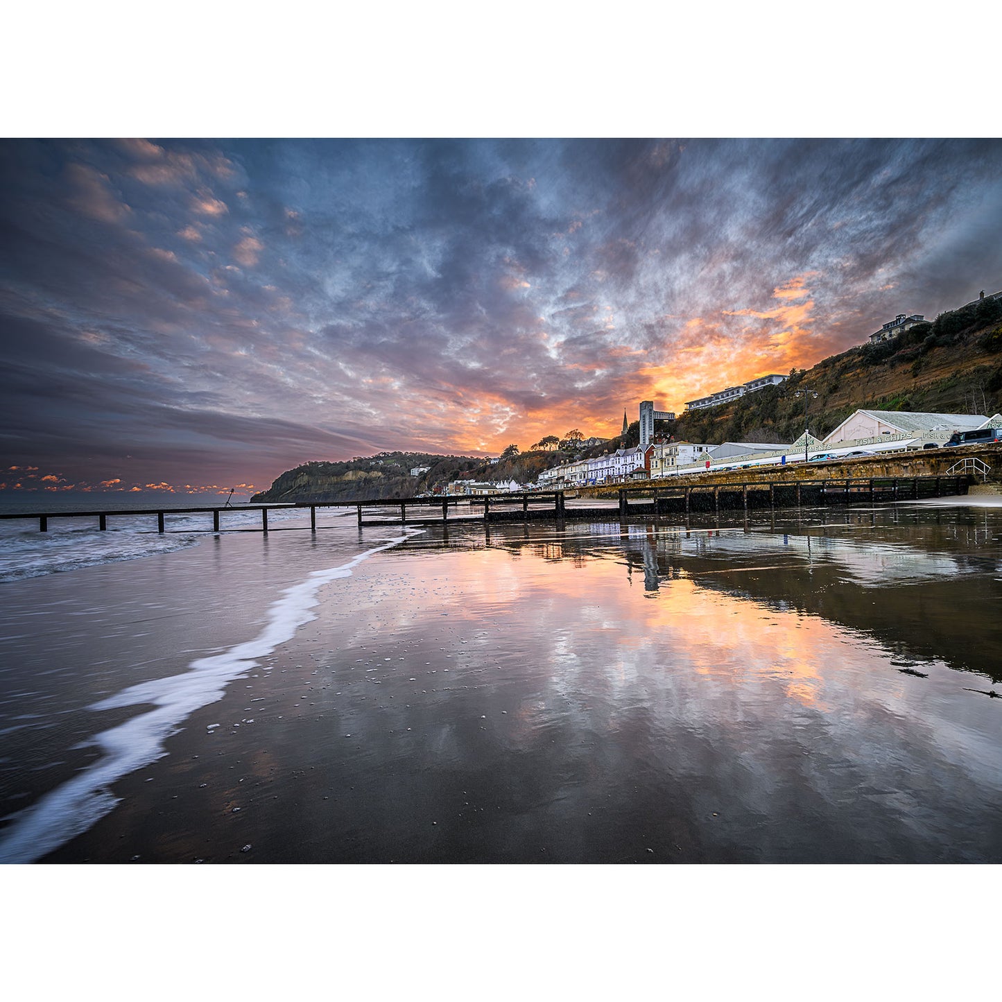 Shanklin Beach by Available Light Photography captures a pier stretching into the sea at sunset, with dramatic clouds, a colorful sky reflecting on wet sand, and distant buildings and hills in the background.