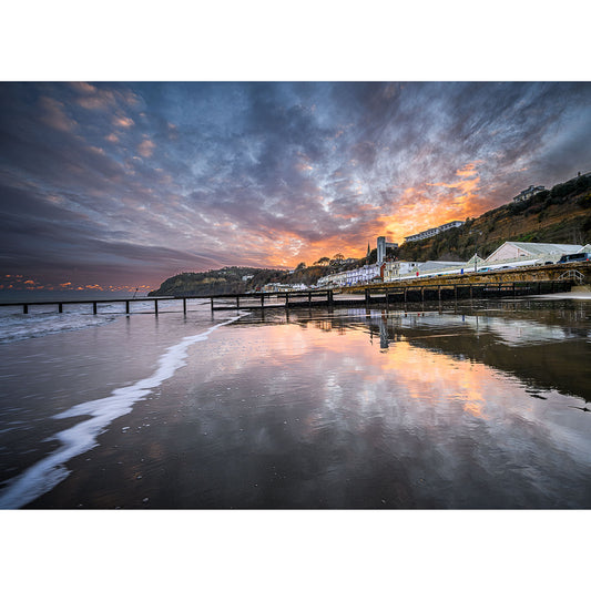 Shanklin Beach by Available Light Photography captures a pier stretching into the sea at sunset, with dramatic clouds, a colorful sky reflecting on wet sand, and distant buildings and hills in the background.