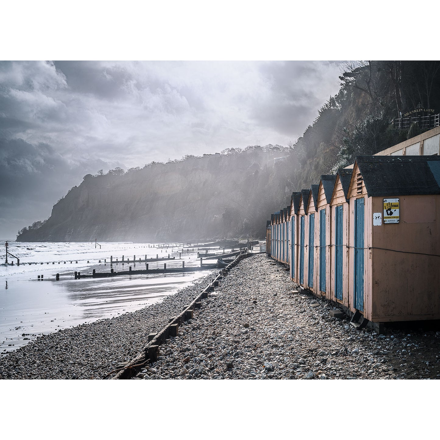 Shanklin Beach by Available Light Photography features a row of colorful beach huts on a pebbled shore beside the sea, set against dramatic cliffs and a cloudy sky.