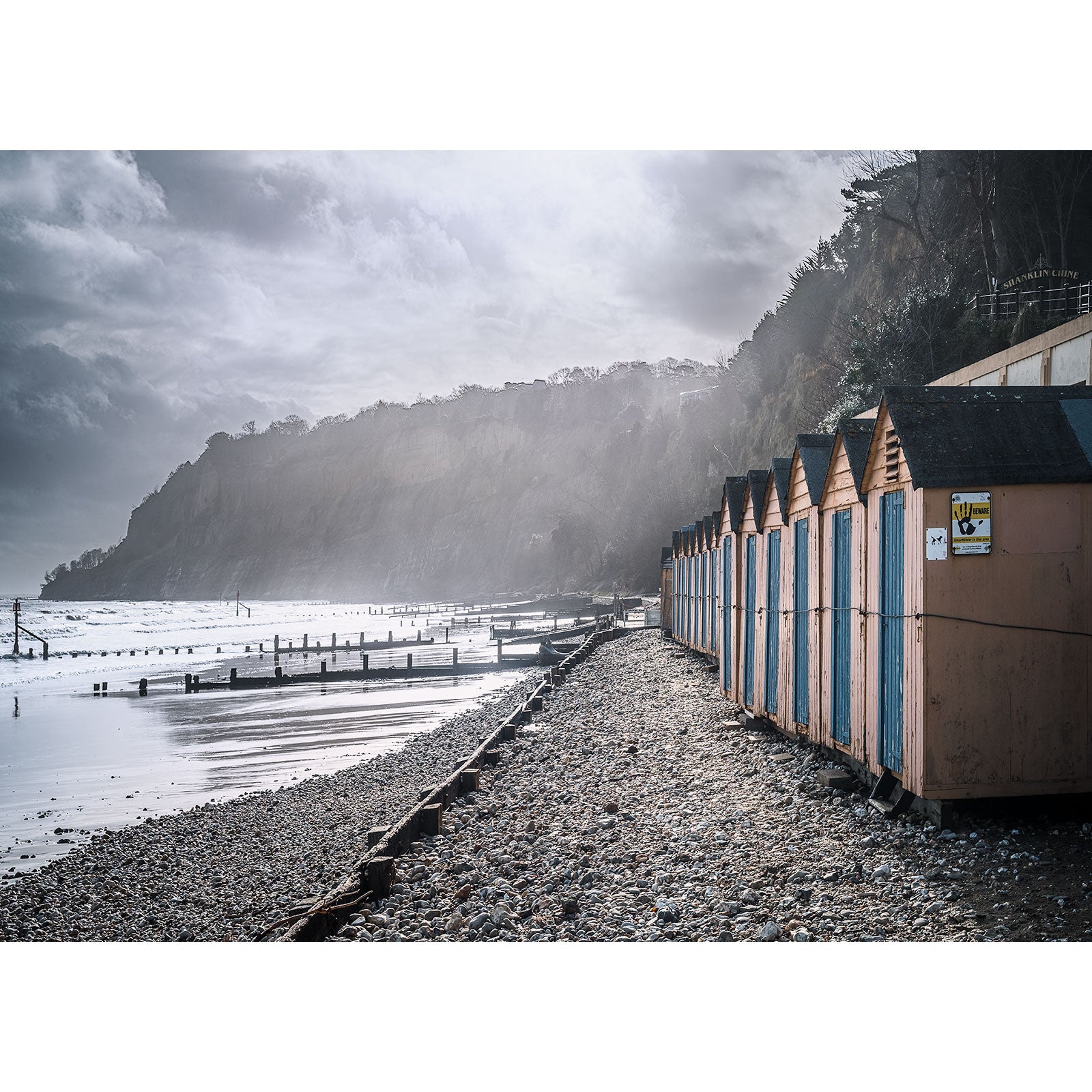 Shanklin Beach by Available Light Photography features a row of colorful beach huts on a pebbled shore beside the sea, set against dramatic cliffs and a cloudy sky.