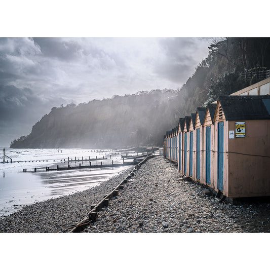Shanklin Beach by Available Light Photography features a row of colorful beach huts on a pebbled shore beside the sea, set against dramatic cliffs and a cloudy sky.