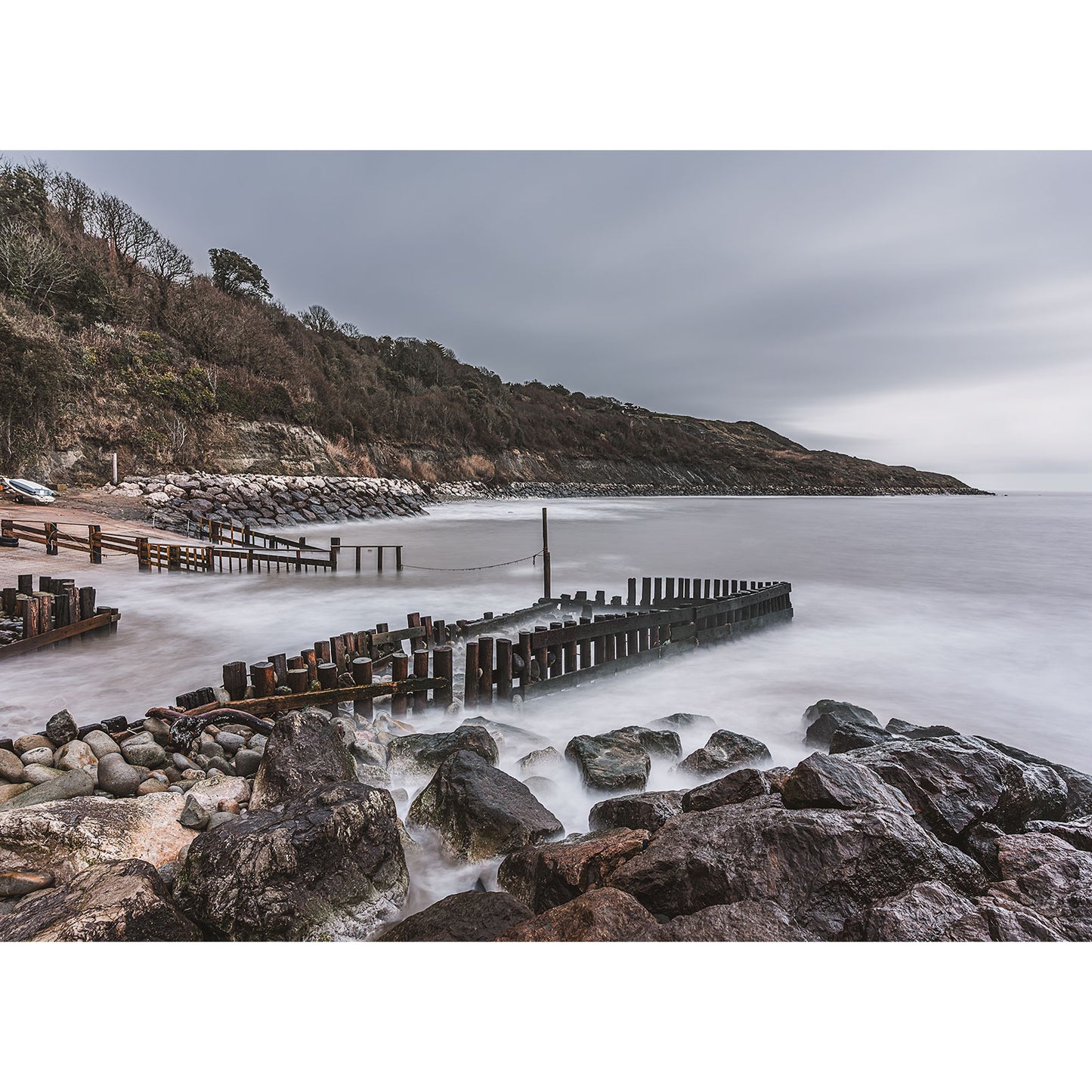 Castlehaven by Available Light Photography captures wooden groynes stretching into calm, misty water, with tranquil hills and trees beneath a cloudy sky—creating a serene, nature-inspired scene.