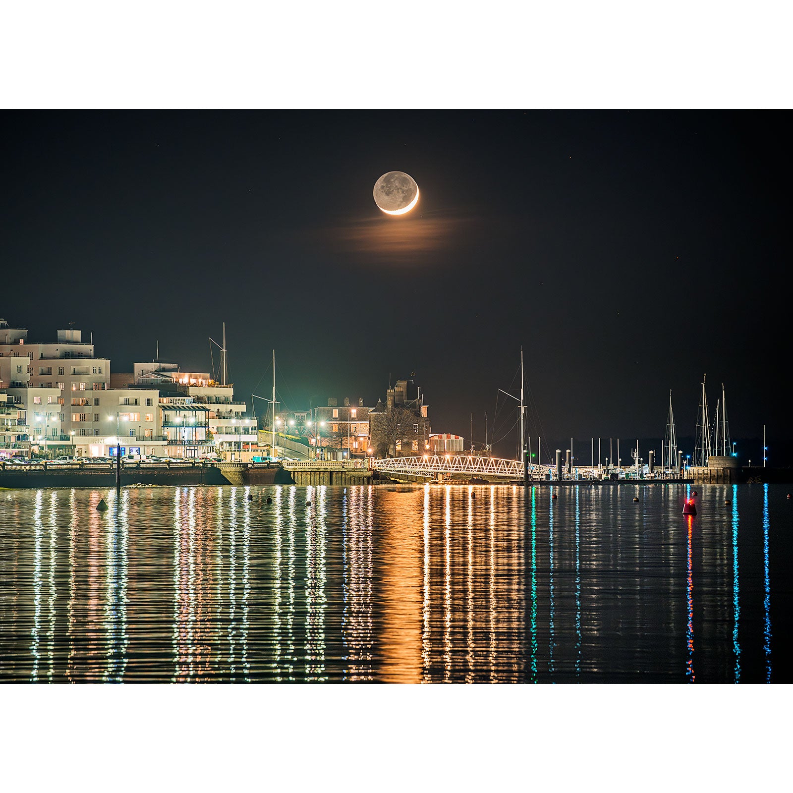 Crescent Moon over Cowes by Available Light Photography captures a low-hanging moon above the harbor, with sailboats gliding past modern buildings and shimmering reflections on calm nighttime waters.