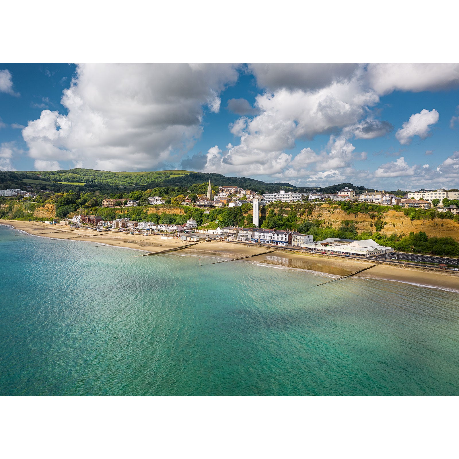 Shanklin by Available Light Photography features a coastal town with sandy beach, calm blue-green waters, and shoreline buildings beneath a partly cloudy sky, with hills and greenery rising in the background.