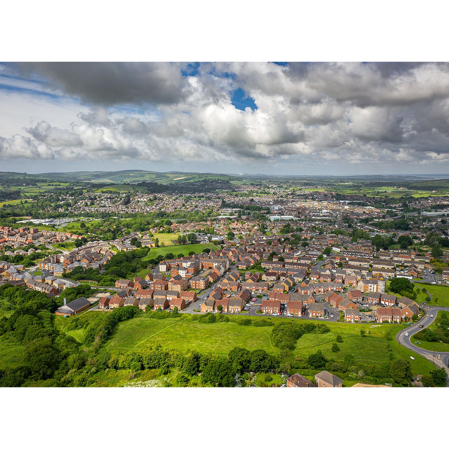 Aerial view of Newport by Available Light Photography showcases residential houses, roads, green fields, and distant hills under a partly cloudy sky.