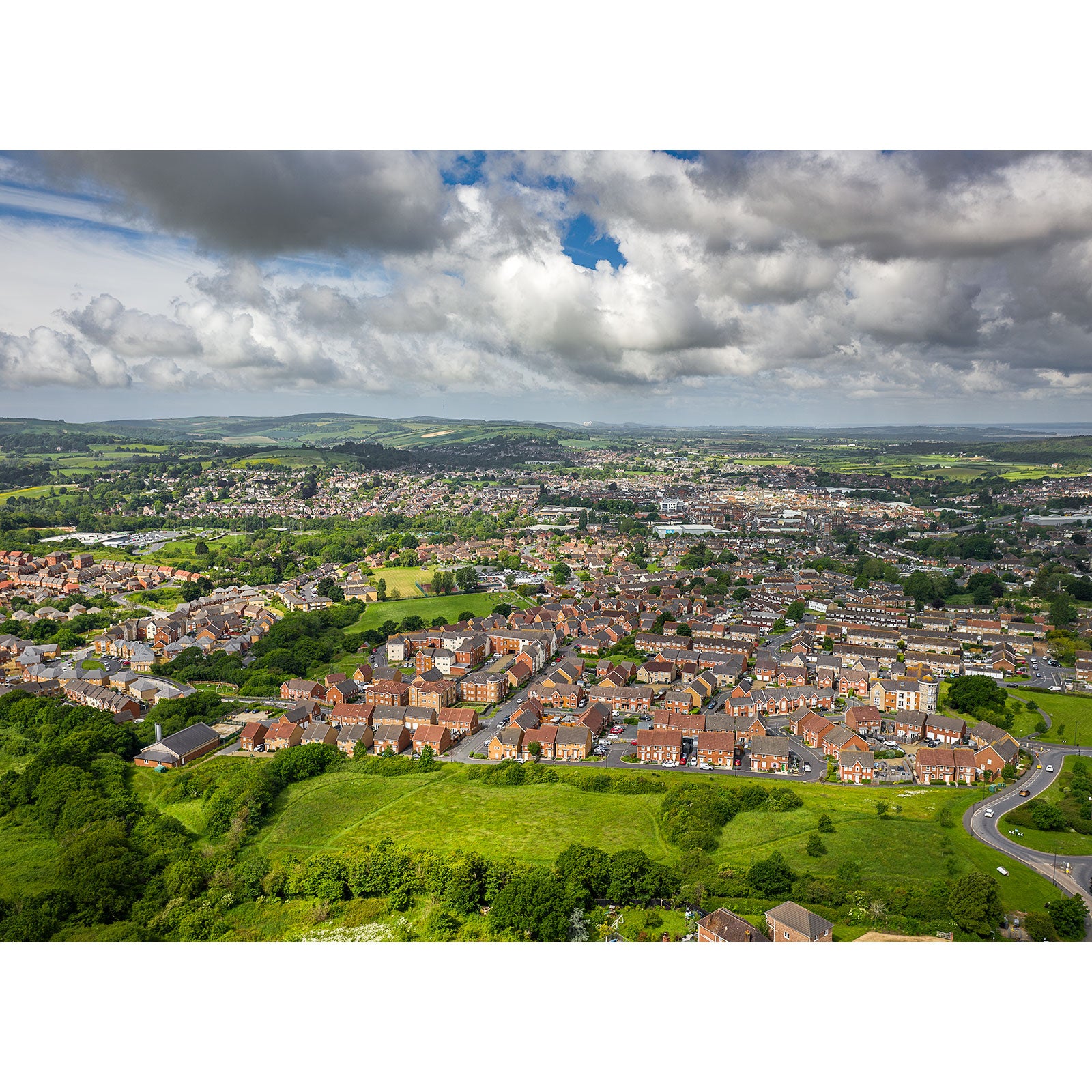 Aerial view of Newport by Available Light Photography showcases residential houses, roads, green fields, and distant hills under a partly cloudy sky.