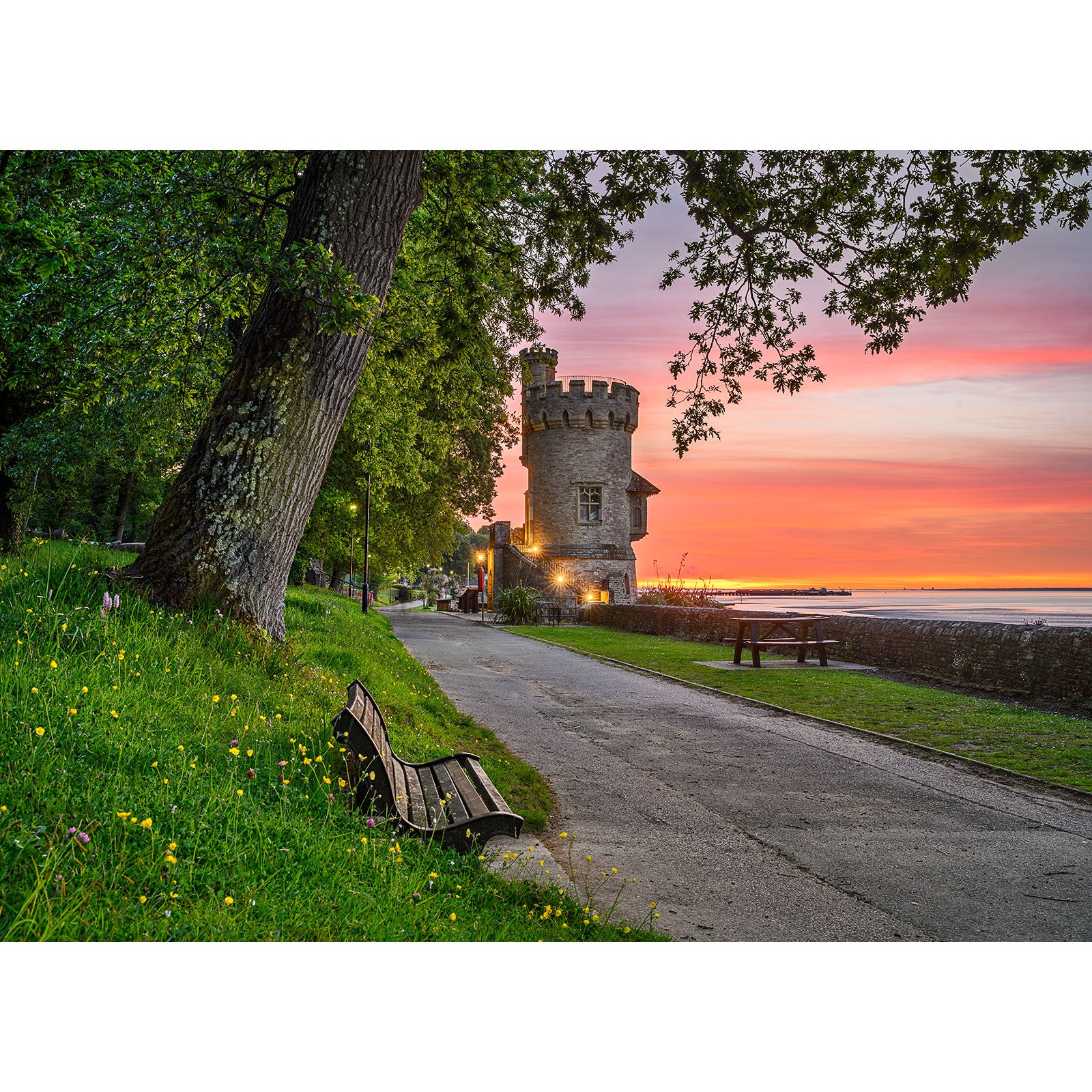 Appley Tower by Available Light Photography captures a stone tower beside a serene waterfront at sunset, with a tree-lined path, benches, and grassy foreground dotted with yellow flowers.