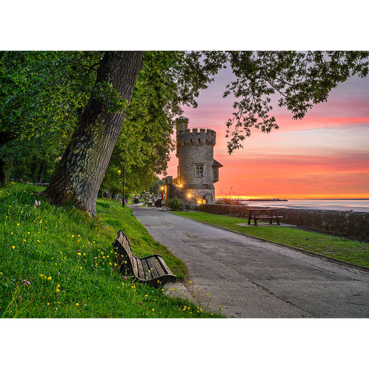 Appley Tower by Available Light Photography captures a stone tower beside a serene waterfront at sunset, with a tree-lined path, benches, and grassy foreground dotted with yellow flowers.