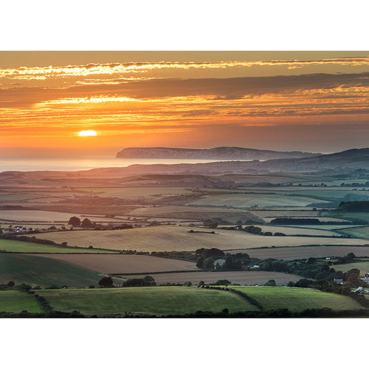 Sunset over a hilly landscape with the sun casting a warm glow over the fields and the distant coastline of Overlooking West Wight, captured by Available Light Photography.