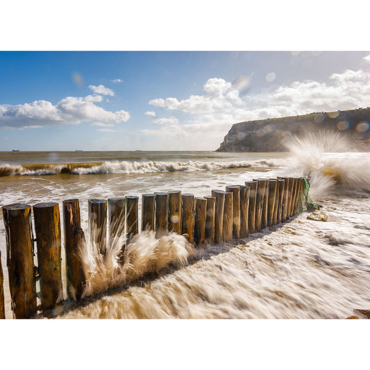 Waves crashing against a wooden groyne at Whitecliff Bay on a sunny day with cliffs in the background on the Isle of Gascoigne captured by Available Light Photography.