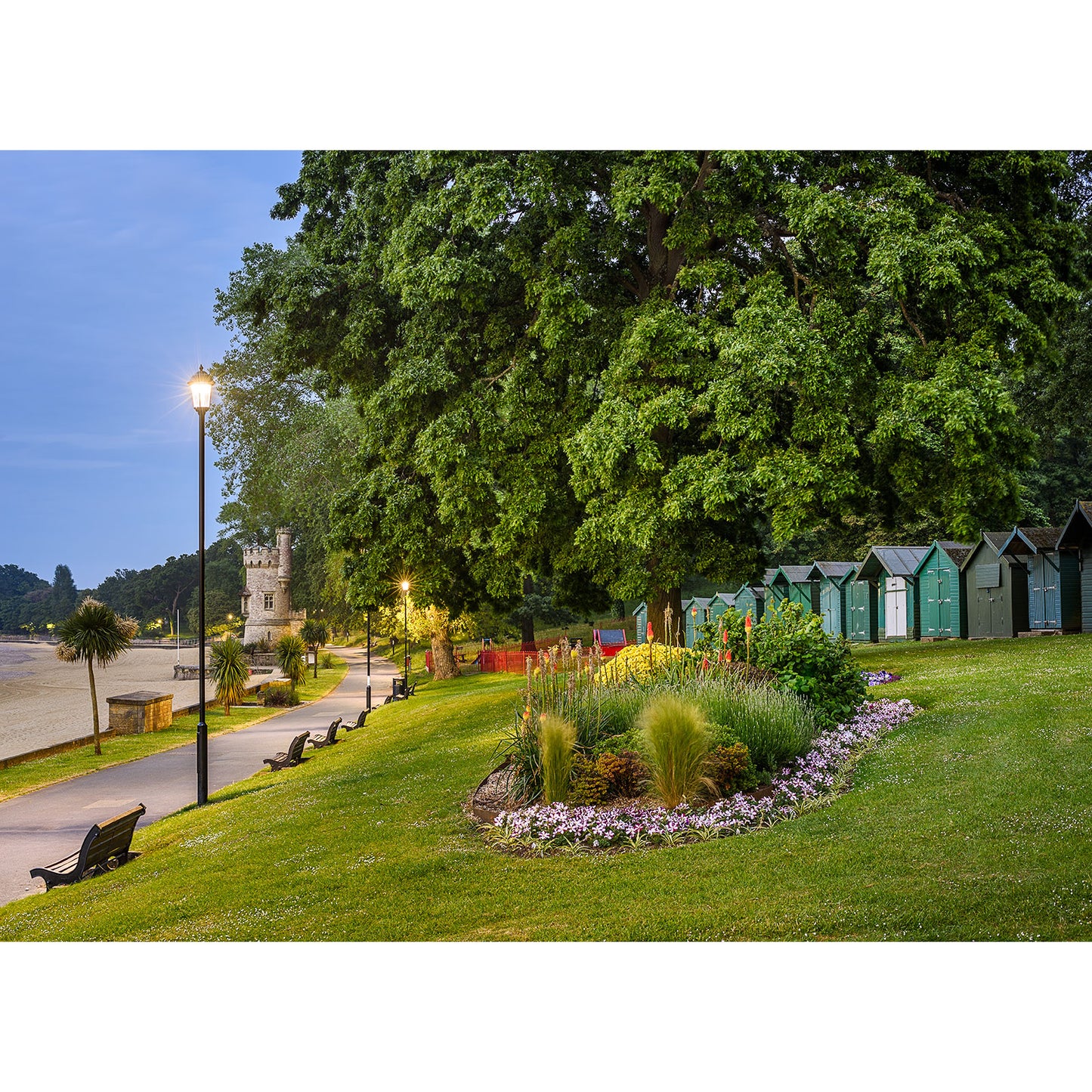 A serene evening in a park with a flowerbed, benches, and colorful beach huts on the Isle of Wight near a sandy area, illuminated by an Appley street lamp from Available Light Photography.