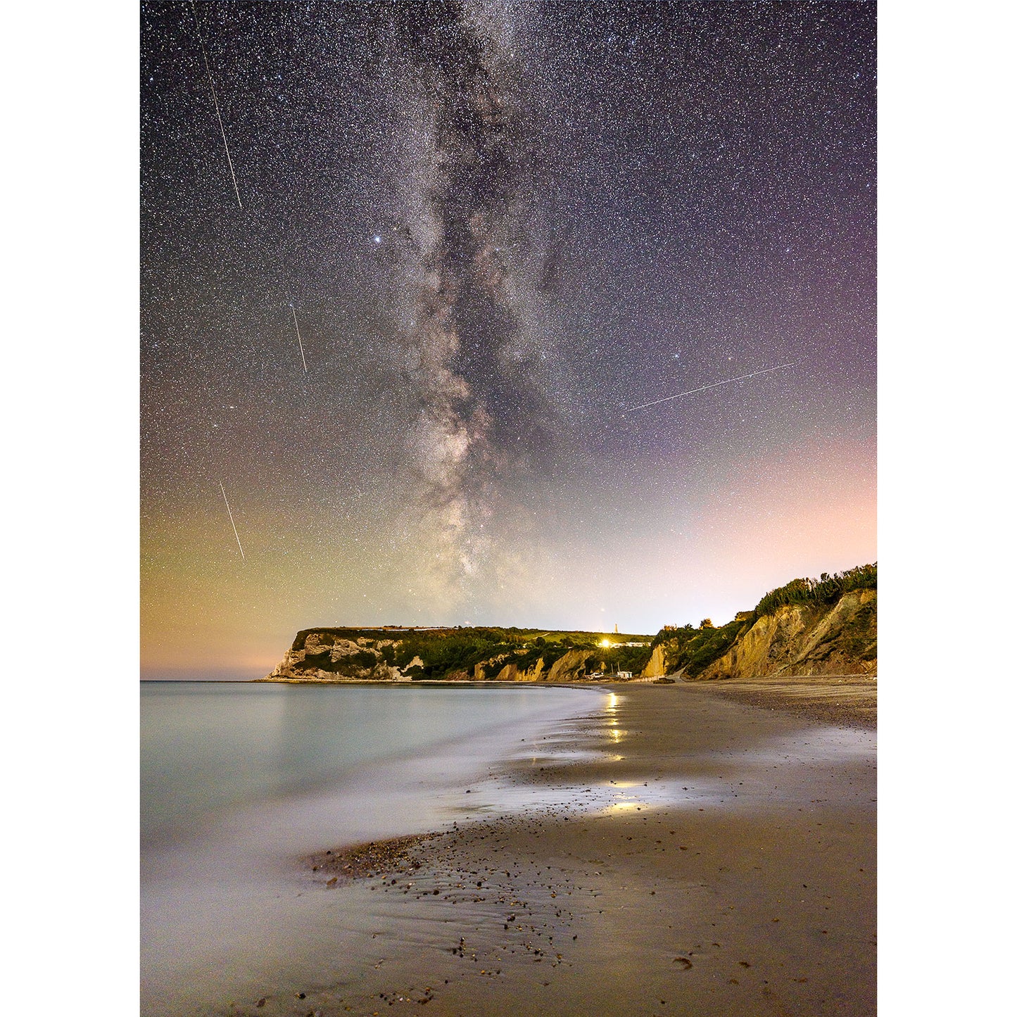 A starry night sky featuring the Milky Way and Perseid Meteors over a tranquil beach on Whitecliff Bay with light reflections on the water's surface by Available Light Photography.