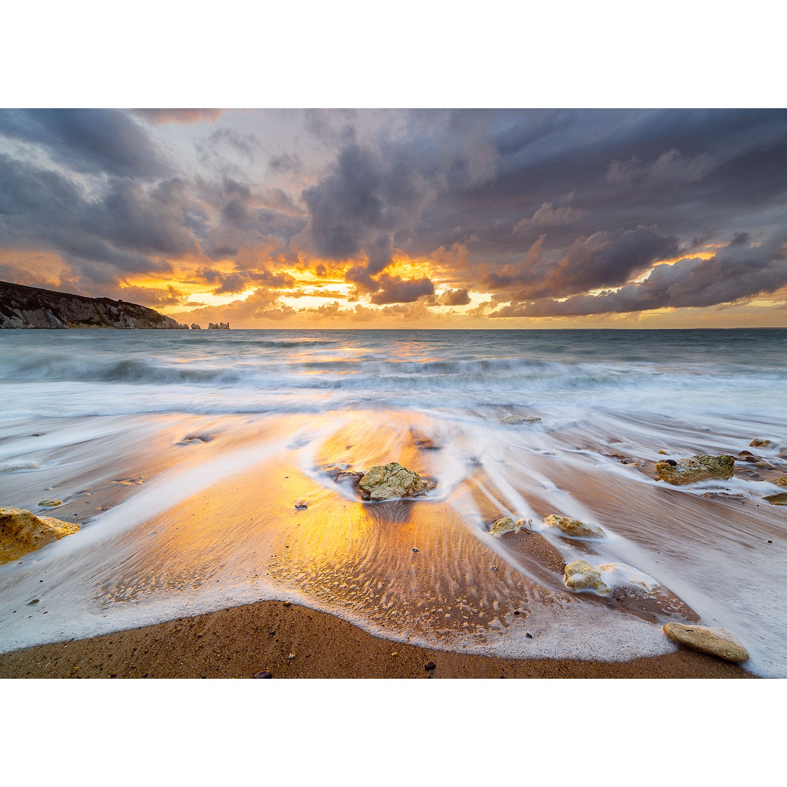Sunset over a turbulent sea with waves washing onto a sandy beach with rocks, under a dramatic cloud-filled sky on the Isle of Wight captured by Alum Bay and The Needles from Available Light Photography.