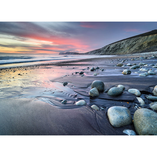 A serene beach at sunset on the Compton Bay, with smooth stones scattered on the sand, highlighted by the vibrant hues of the twilight sky by Available Light Photography.
