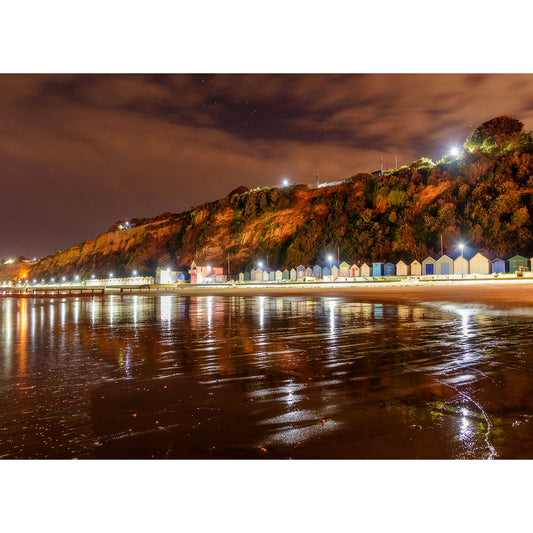 A night view of brightly lit Lake Beach huts along a shoreline with a cliff in the background under a starry sky on the Isle of Wight by Available Light Photography.