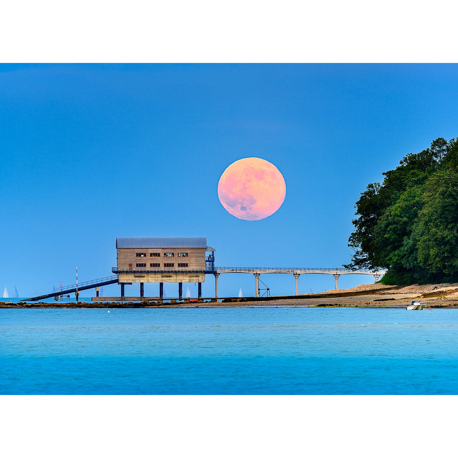 A large, pink-toned Moonrise rising over a coastal structure connected by a pier on Gascoigne Isle, with clear blue skies and trees in the background, captured by Available Light Photography.