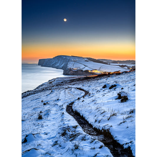 A winding path through a snowy landscape leading to cliffs by the Isle of Wight under a twilight sky with a visible Crescent Moon, Tennyson Down captured by Available Light Photography.