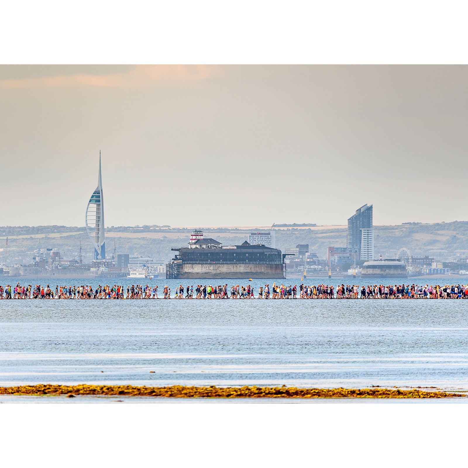 A large group of people, including Steve, walking along the shoreline on the Isle of Wight with a city skyline in the background captured by the Available Light Photography's The Fort Walk.