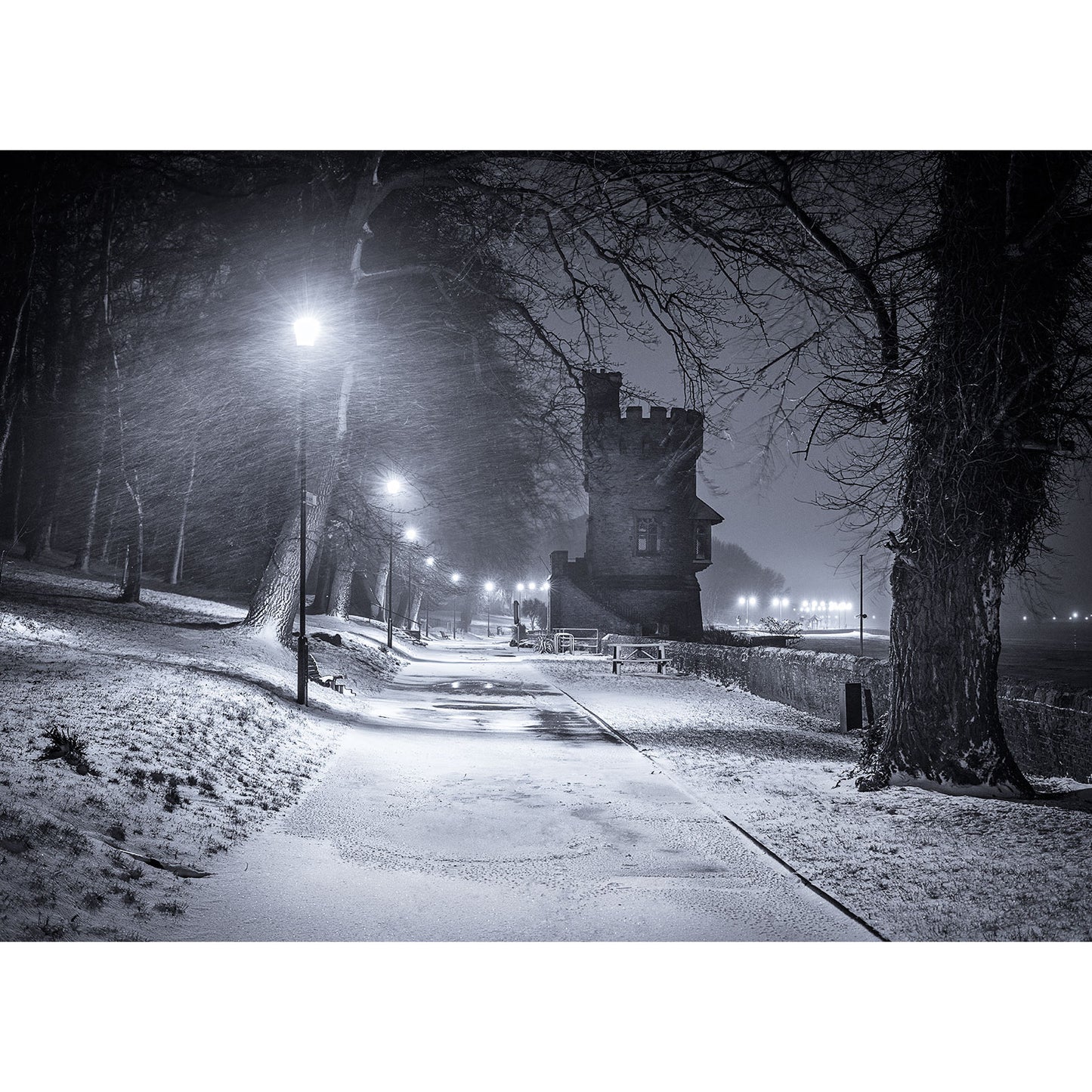 A snowy night scene with a lamp-lit pathway leading to an old tower on the Isle of Wight, flanked by bare trees and a glowing cityscape in the distance captured by Winter's Night, Appley Tower from Available Light Photography.