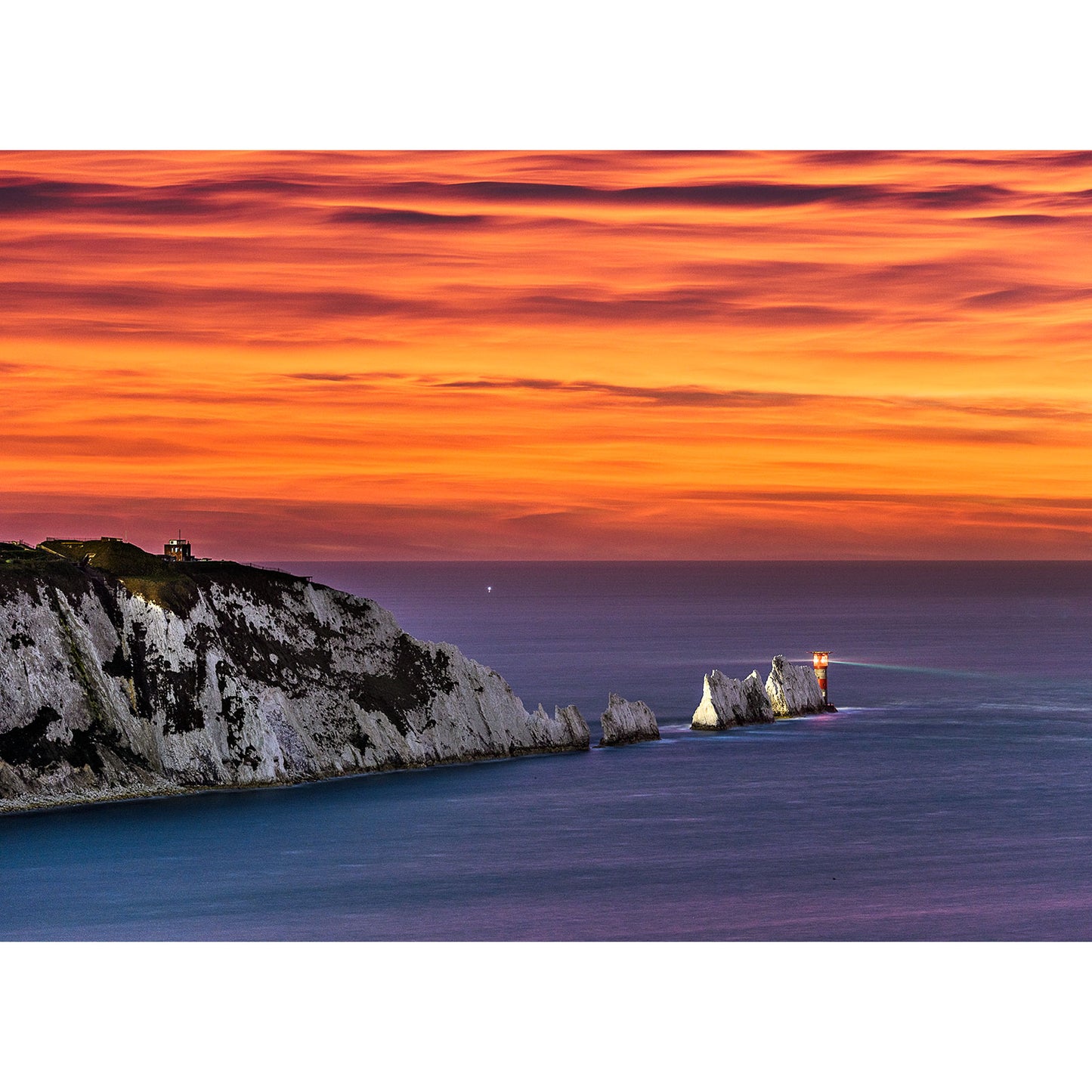A vibrant sunset over the Isle of Wight's coastal landscape with white cliffs and a lighthouse captured in "The Needles by Moonlight" by Available Light Photography.