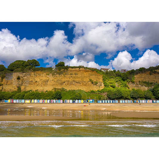 Colorful beach huts lining the shore beneath a tall cliff against a backdrop of blue sky and fluffy clouds on Hope Beach, Shanklin by Available Light Photography.
