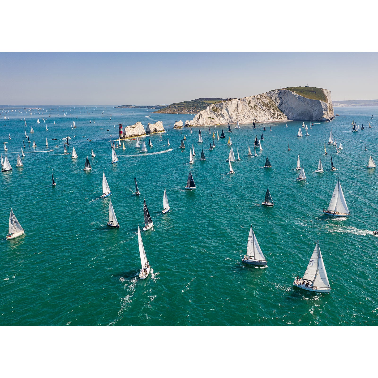 A fleet of Round the Island Race sailboats racing near a coastal cliff on the Isle of Wight on a clear day, captured by Available Light Photography.
