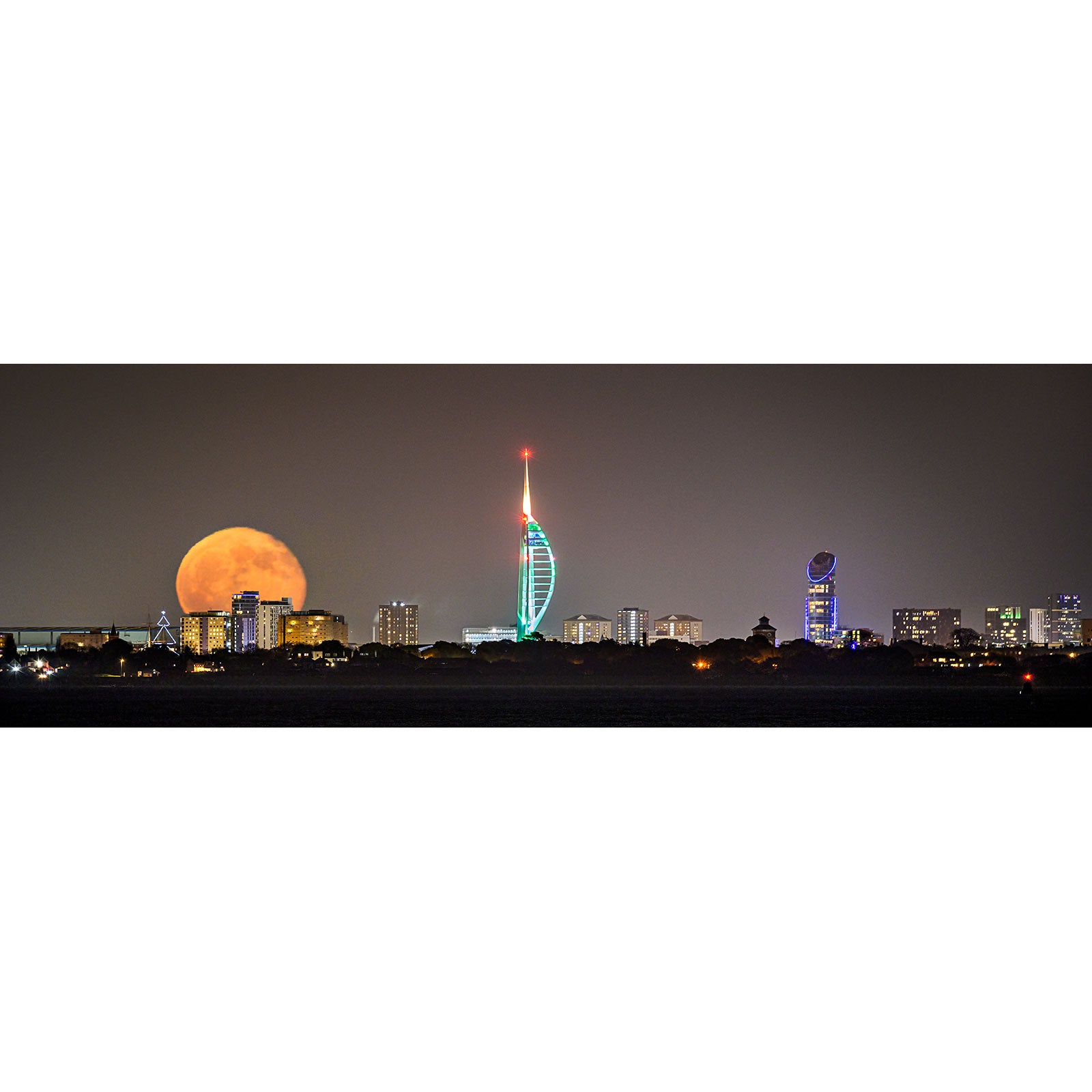 A Moonrise Spinnaker Tower rising behind a city skyline on the Isle of Wight, with an illuminated skyscraper at night. By Available Light Photography.