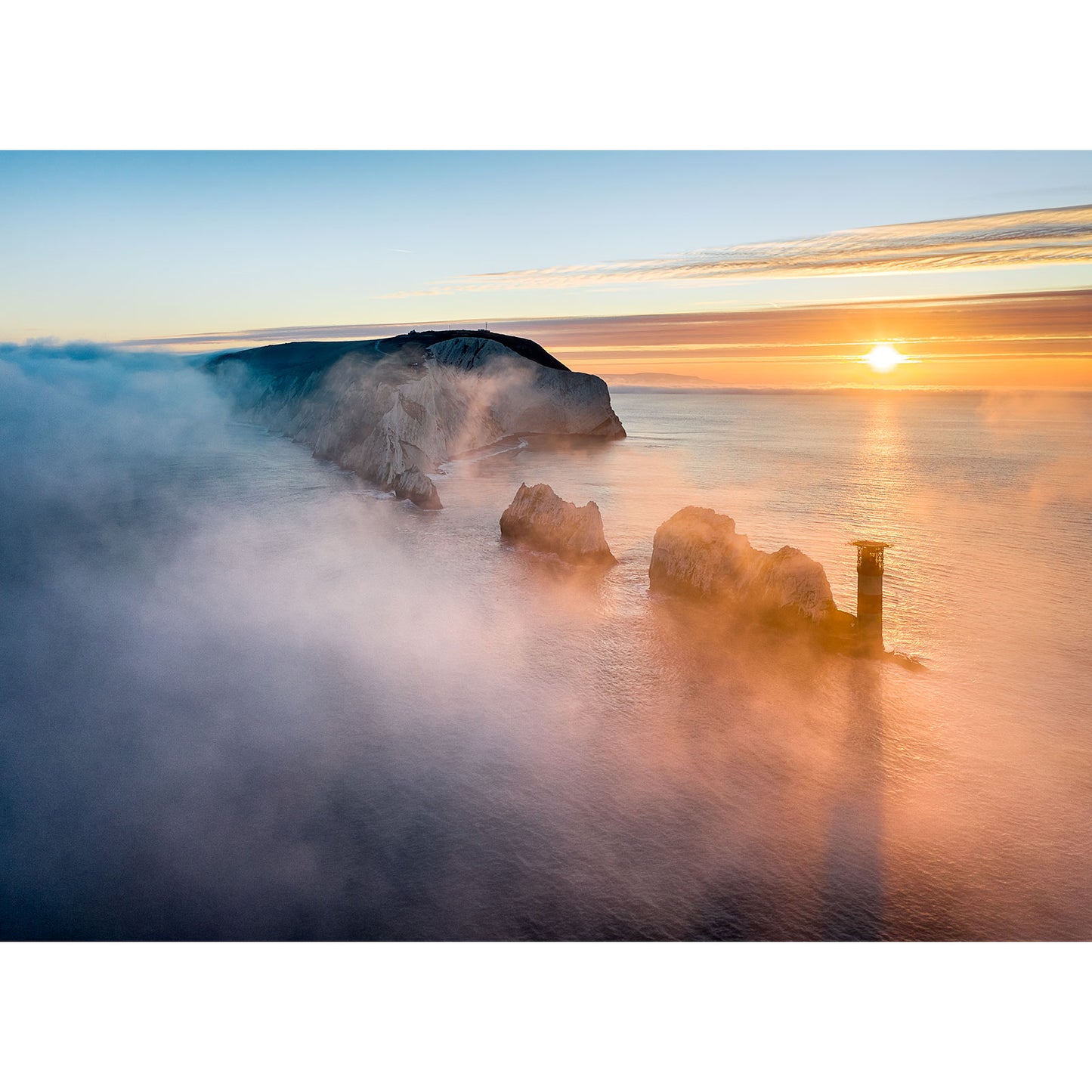 Sunrise over a misty coastline with rock formations and a lighthouse on the Isle of Fog at The Needles by Available Light Photography.