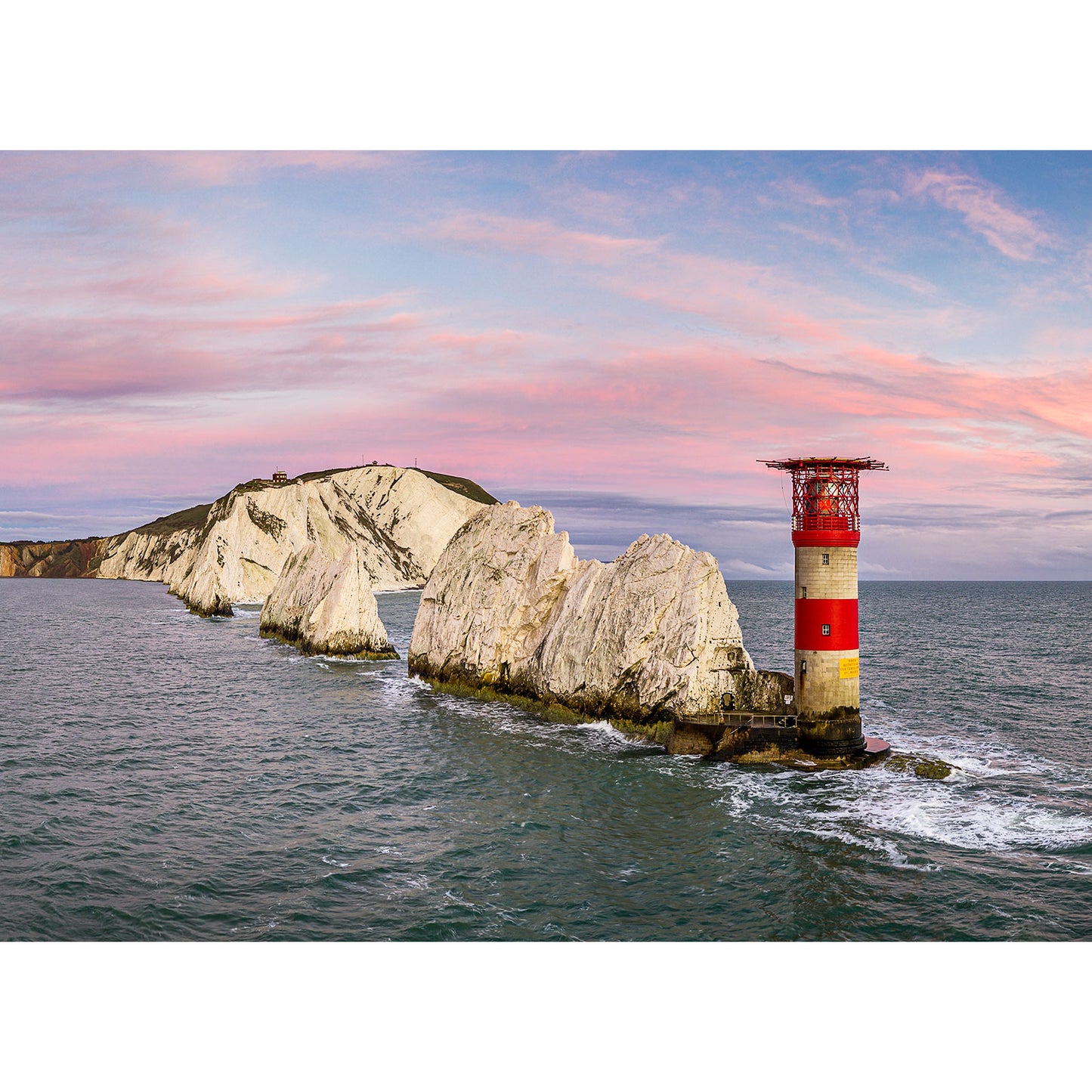 A "The Needles" lighthouse stands on a rocky outcrop near a coastal cliff on the Isle of Wight under a soft pink sky by Available Light Photography.