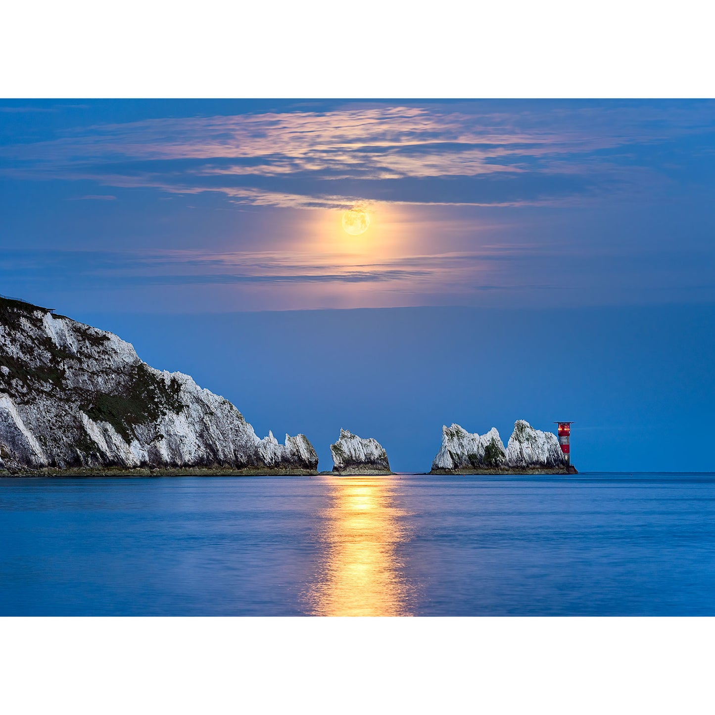A Moonset over The Needles rises above a calm sea, casting a golden reflection on the water, with a lighthouse and white cliffs on the Isle of Wight captured by Available Light Photography.