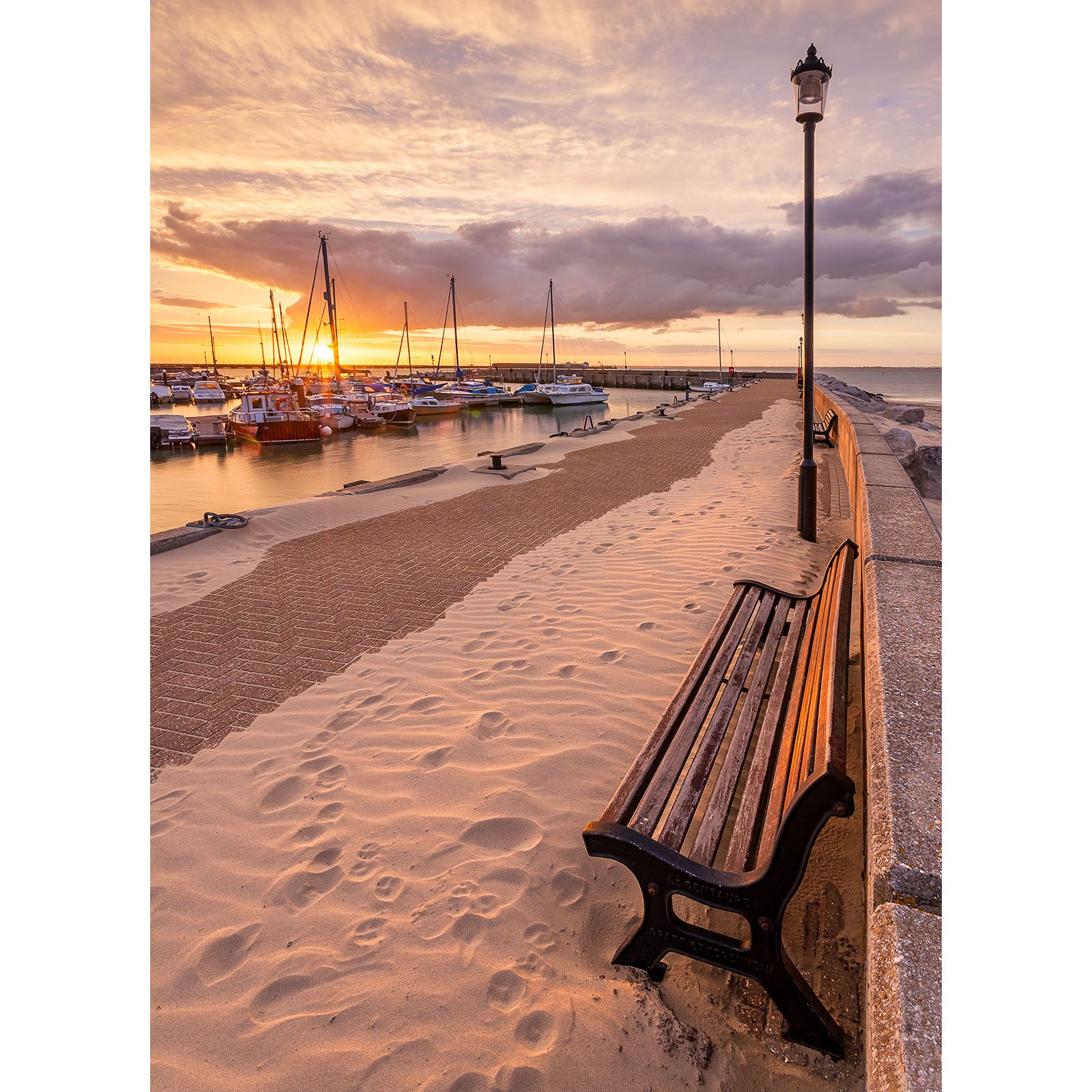 Sunset at Ryde Harbour with boats, a bench, and sand-covered pavement on the Isle of Wight by Available Light Photography.