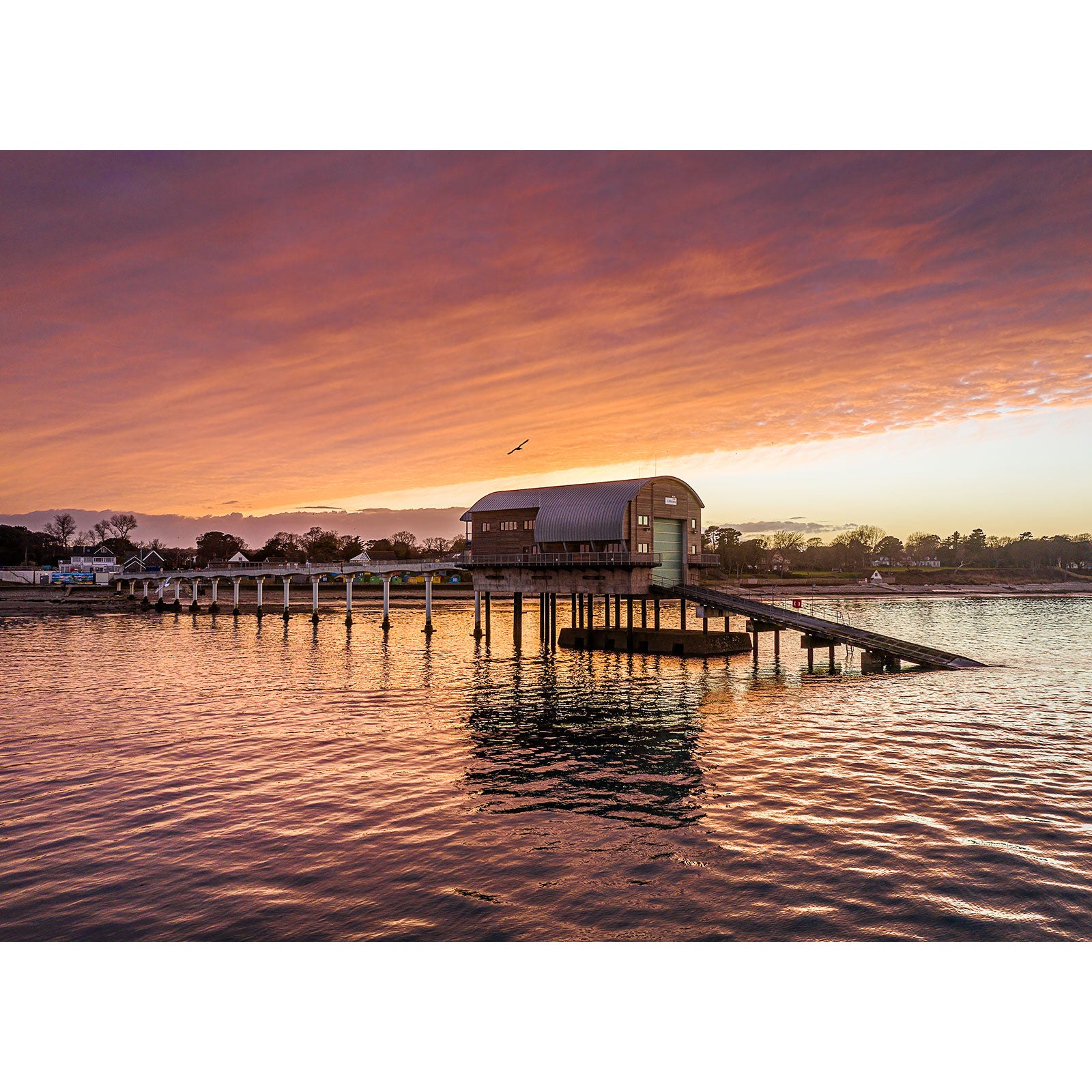 Sunset over a tranquil sea with a pier leading to a building on stilts, reminiscent of the landscapes Available Light Photography often photographed featuring the Bembridge Lifeboat Station.