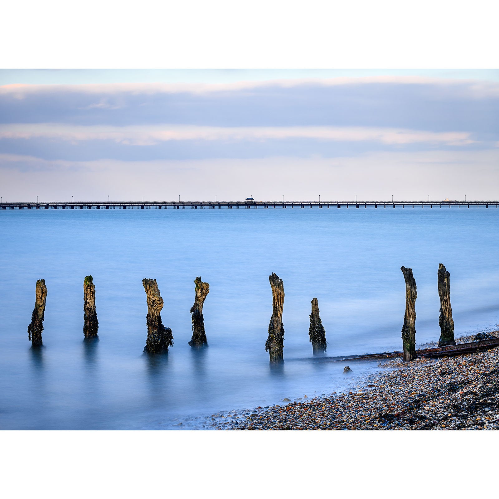 Old wooden pilings on a pebbly beach at the Isle of Wight with Ryde Pier in the background under a blue sky at dusk. (Photography by Available Light Photography)
