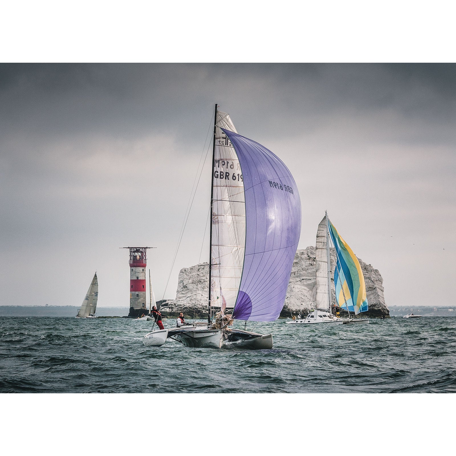 Sailboats racing near the Isle of Wight coastline with a lighthouse and rock formation in the background captured by Available Light Photography's Round the Island Race.