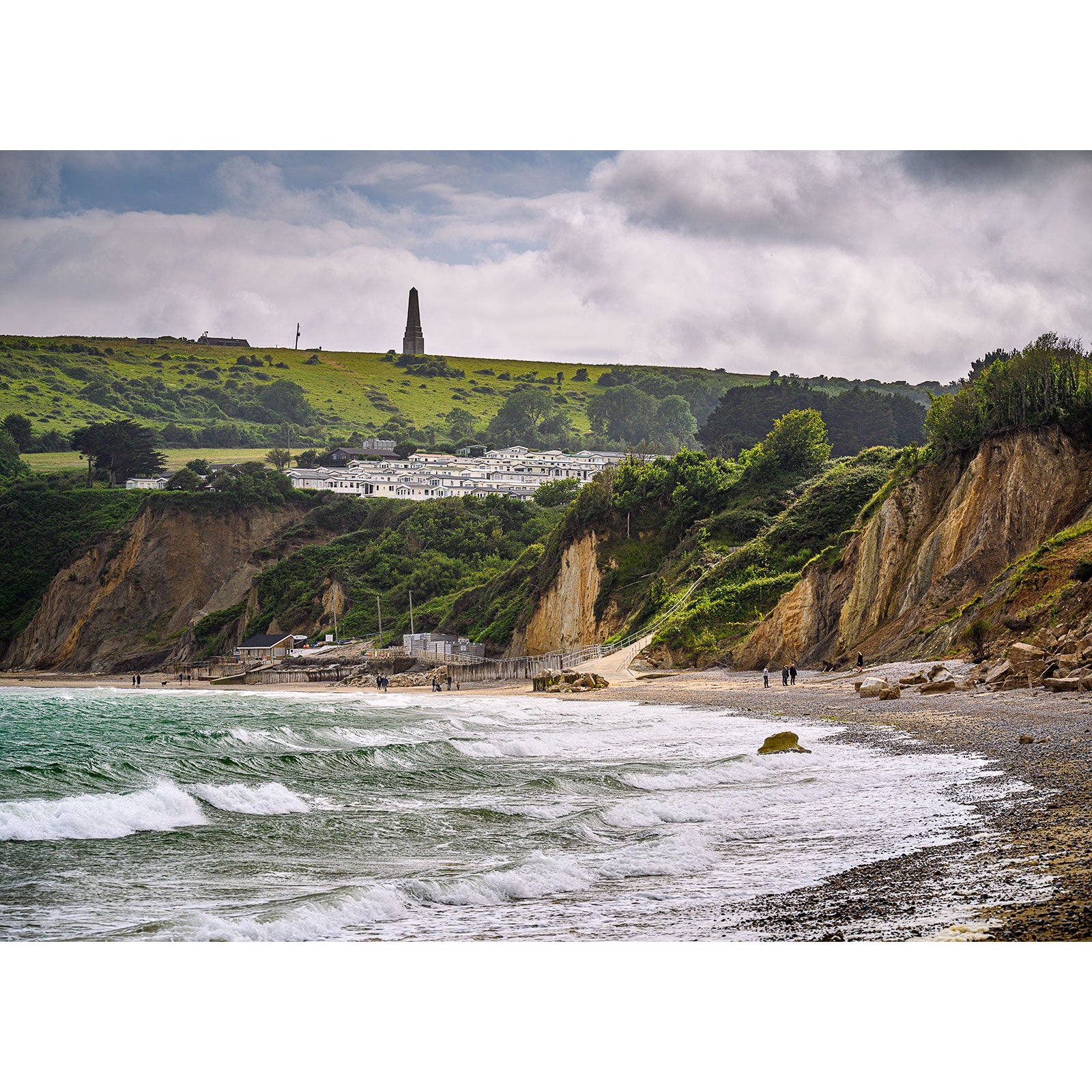 Coastal landscape with cliffs, a pebble beach, and a village near a monument on a hill under a cloudy sky at Whitecliff Bay on the Isle of Wight by Available Light Photography.
