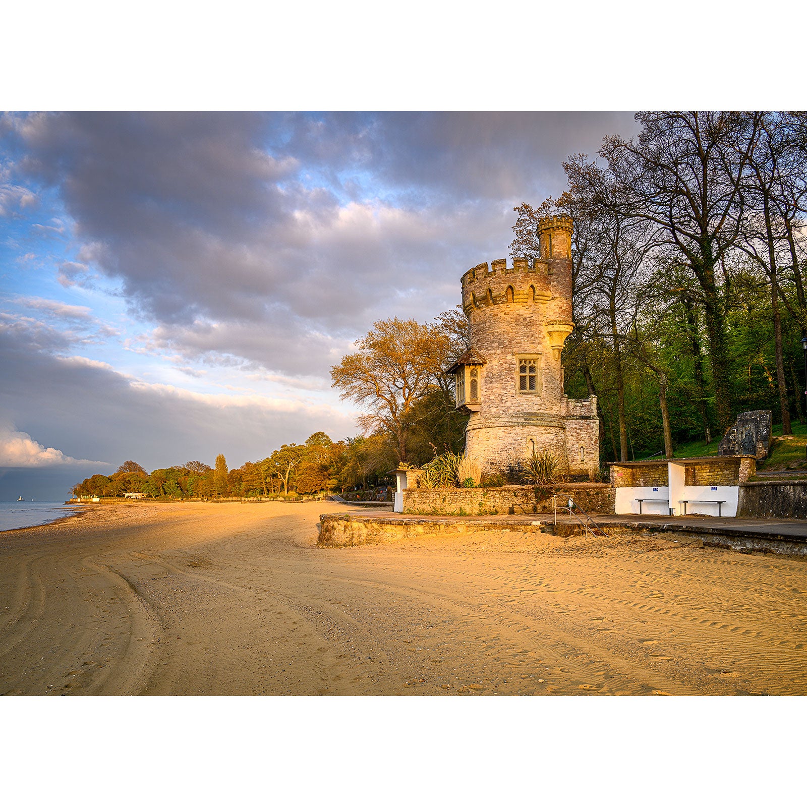 A historic Appley Tower stands adjacent to a sandy beach on the Isle of Wight with a backdrop of trees under a partly cloudy sky.