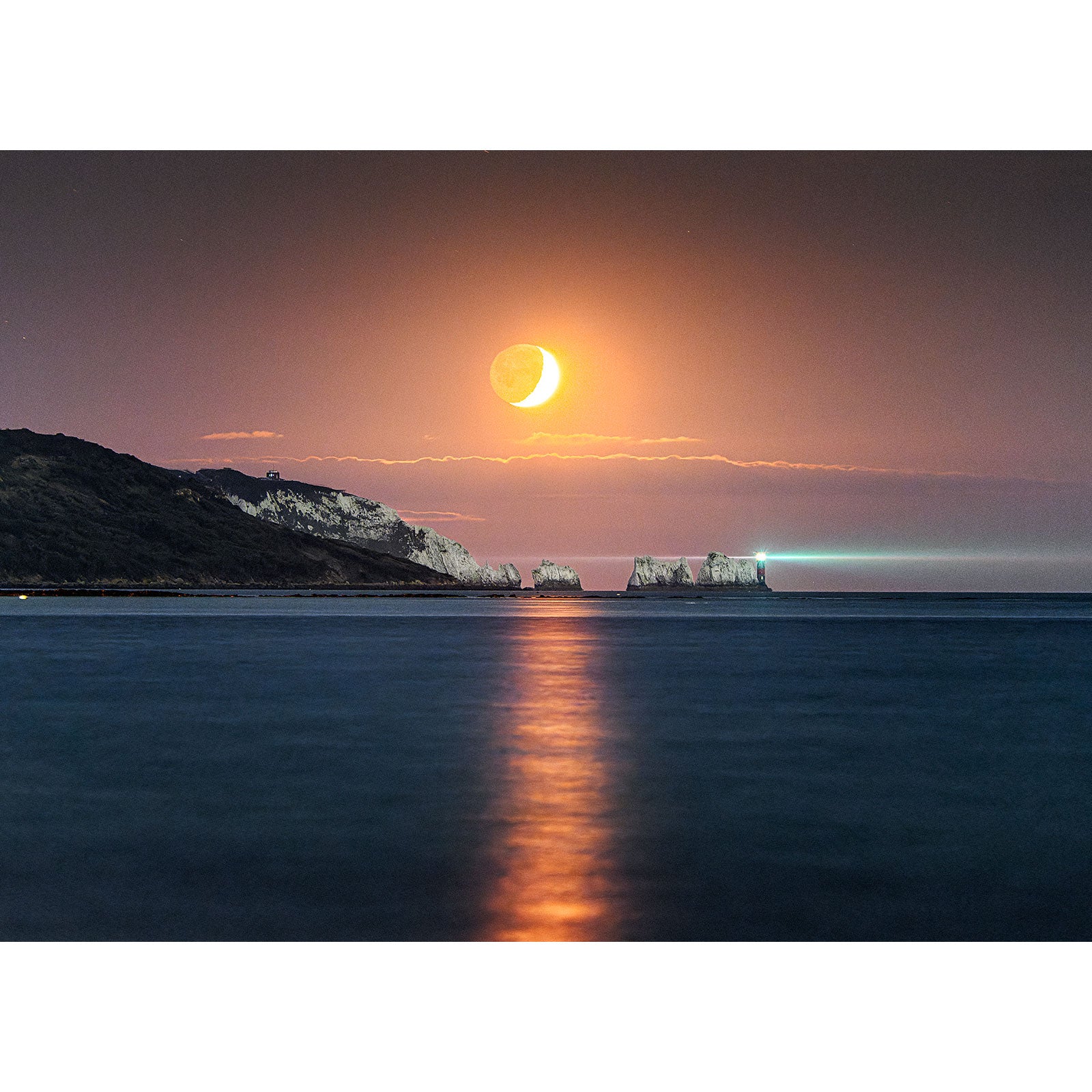 Crescent Moonset at The Needles over a calm sea with a cliff coastline in the distance on the Isle of Wight by Available Light Photography.